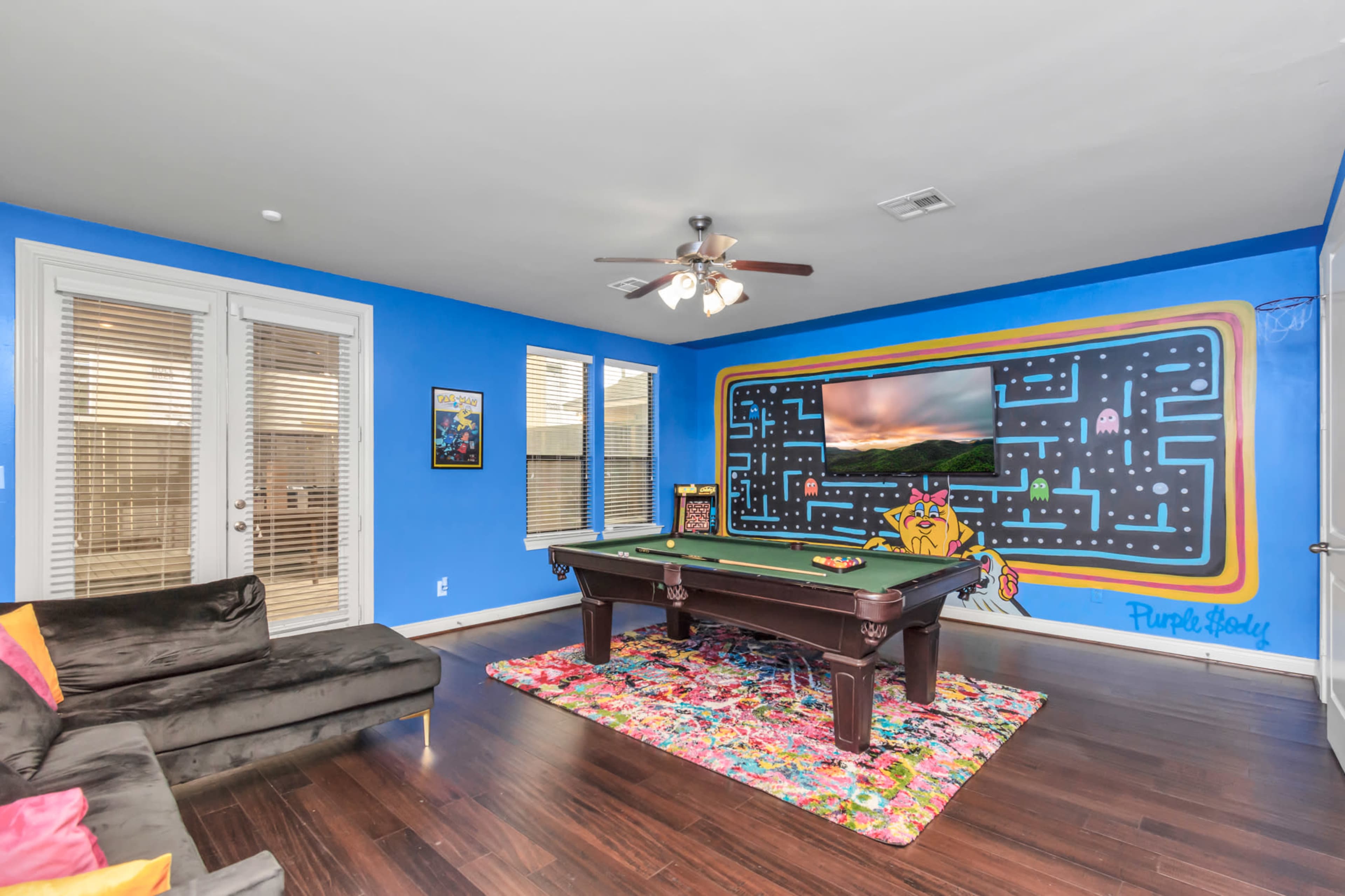 The image shows a game room with a pool table, a colorful rug, a large television on the wall, and blue painted walls featuring a mural.
