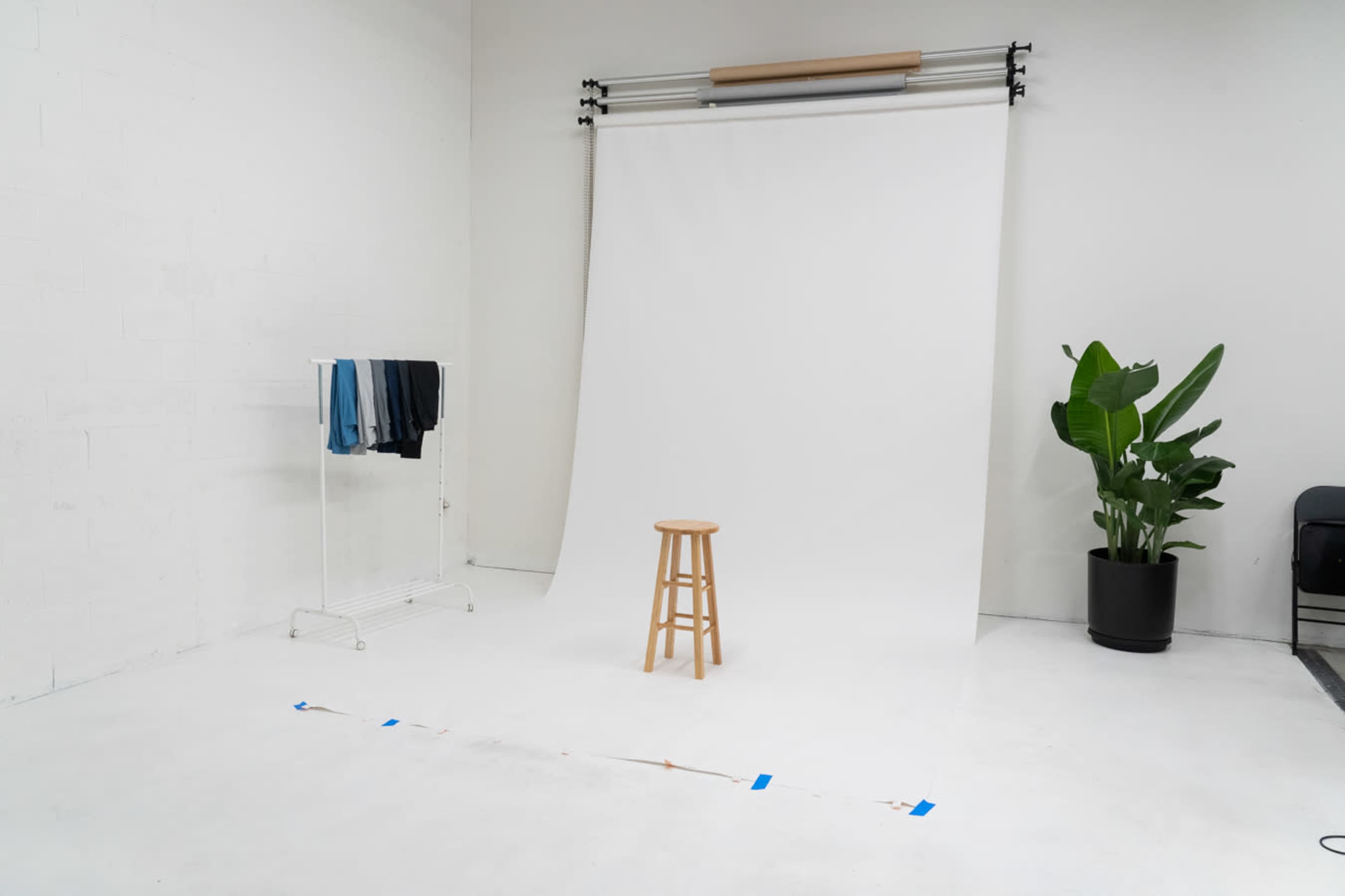 A minimalist photography studio with a white backdrop, a wooden stool at the center, a clothing rack with folded garments, and a potted plant in a corner.
