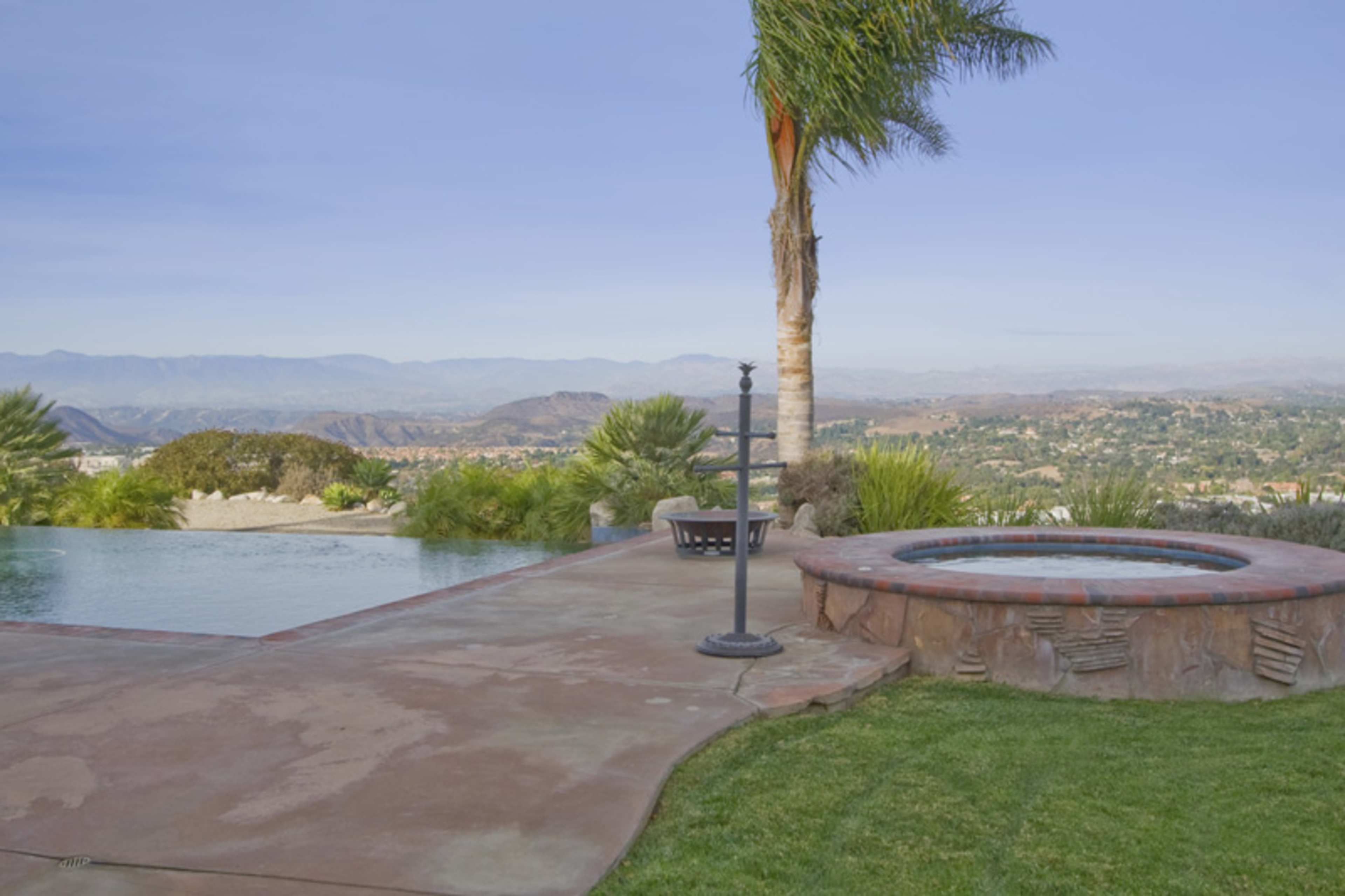 A backyard scene features a swimming pool adjacent to a hot tub, with a view of rolling hills and a clear sky in the background.