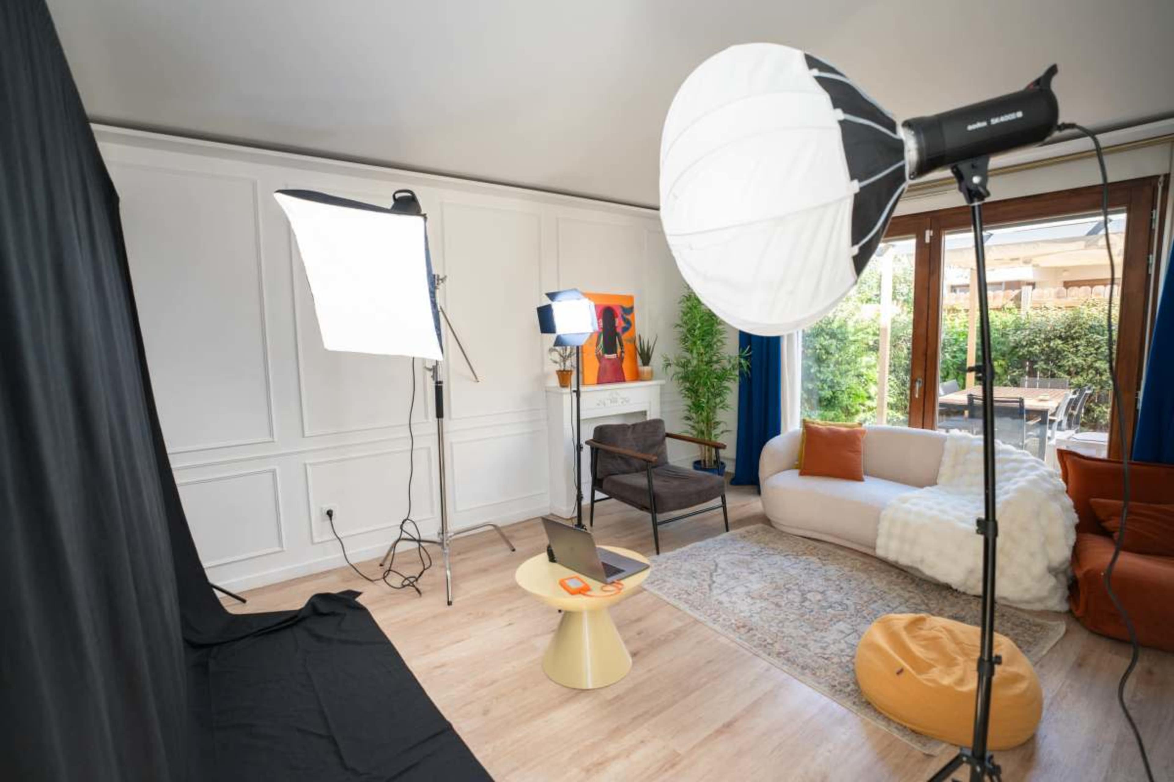 The image shows a well-lit room designed for photography or video production, featuring various lighting equipment, a backdrop, and seating arrangements.