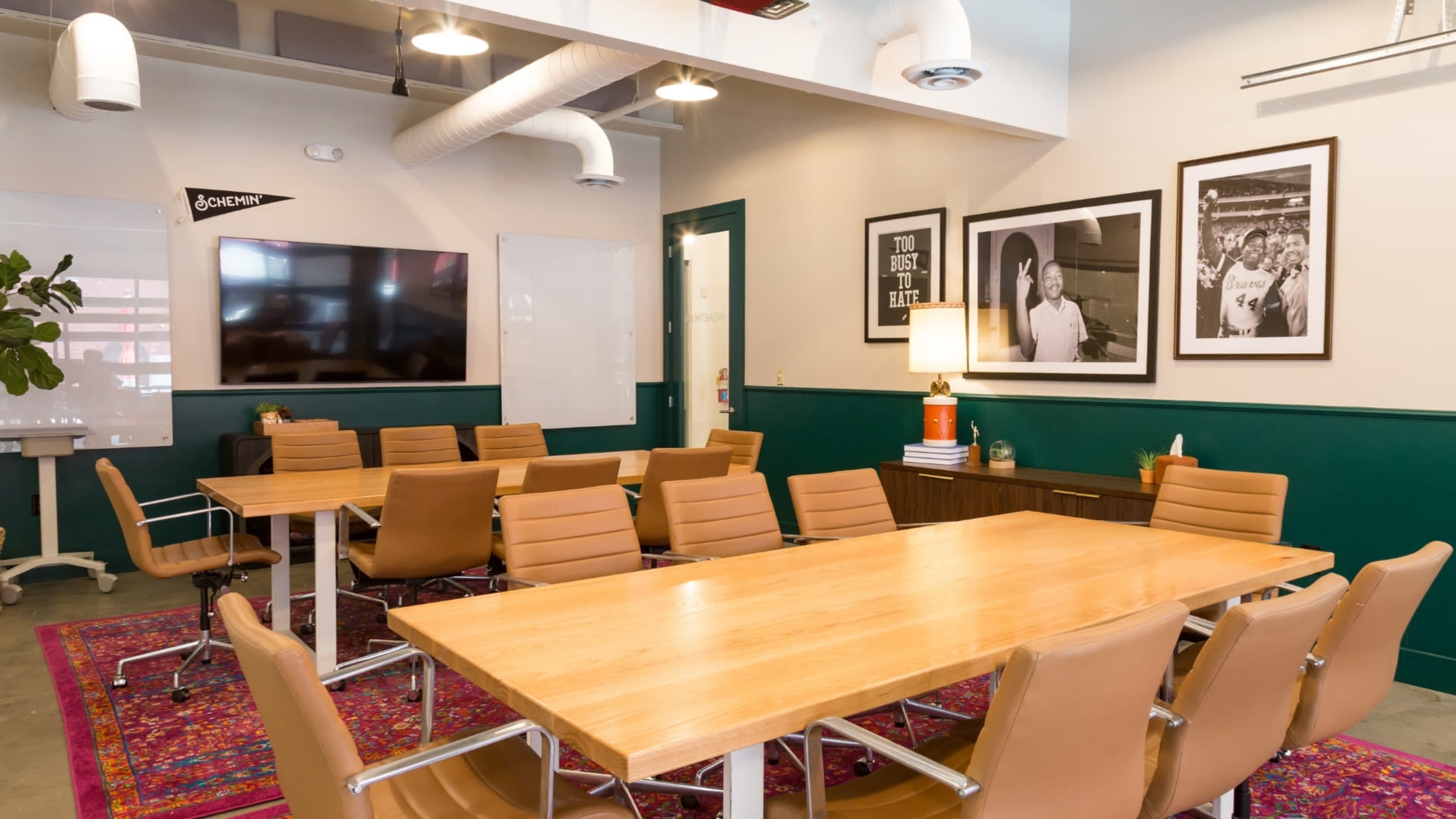 A modern conference room features a large wooden table surrounded by brown leather chairs, with a TV screen and framed photographs on the walls.