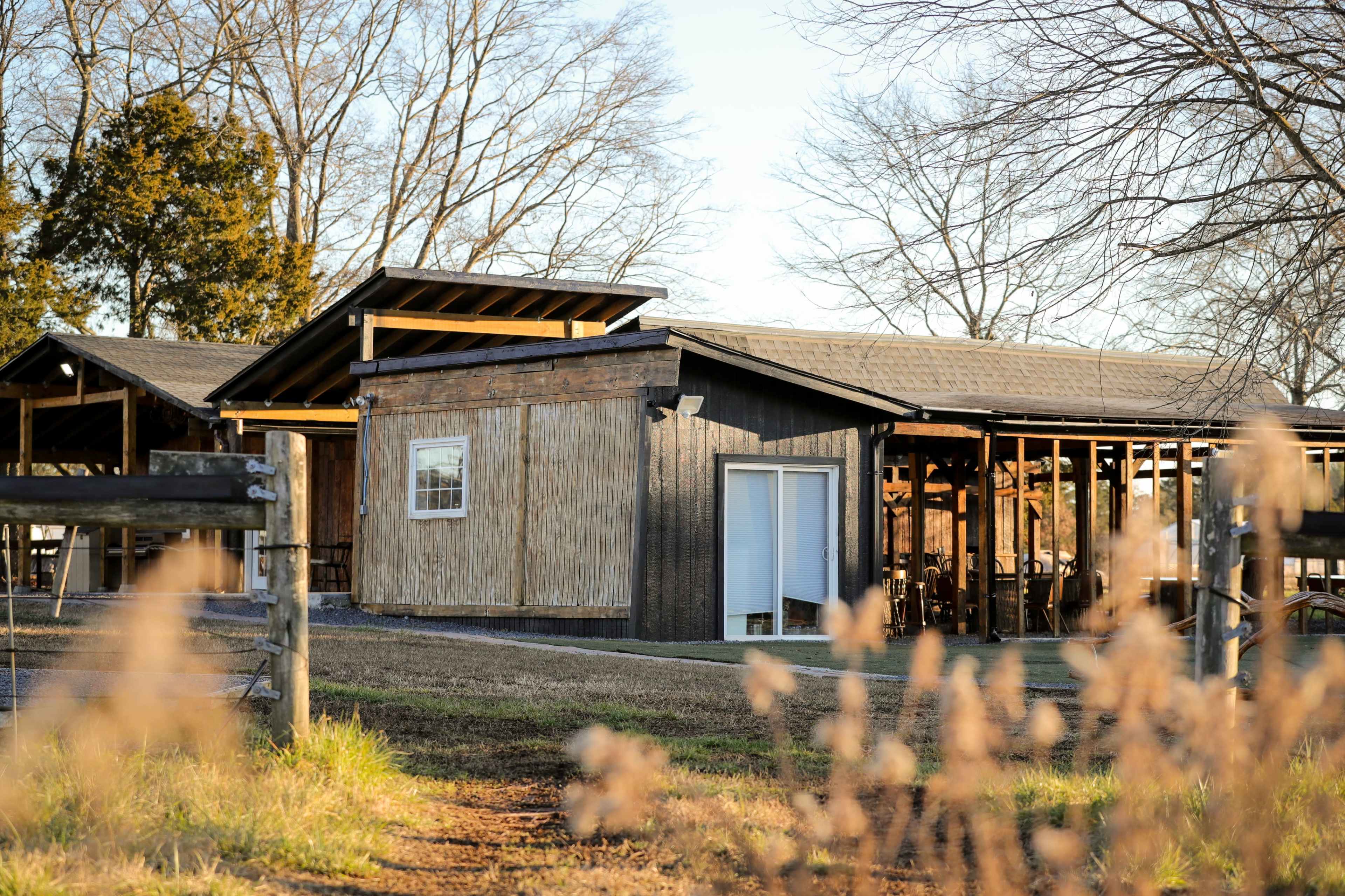 A modern wooden structure with large windows is situated among bare trees and grass, featuring a wraparound porch and nearby fencing.