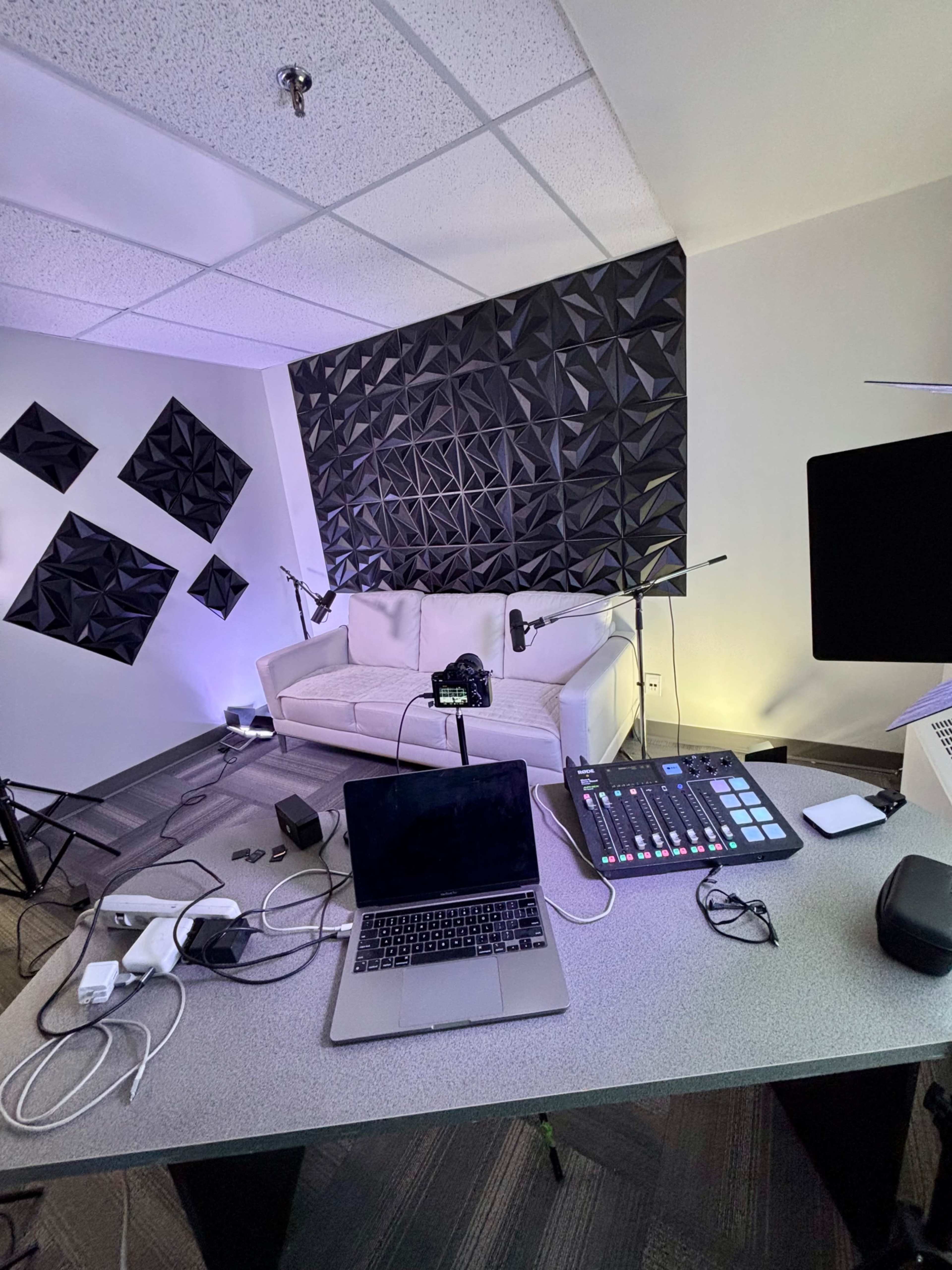 The image shows a modern recording studio with a couch, multiple microphones set up, and a laptop on a table surrounded by various cables and equipment.