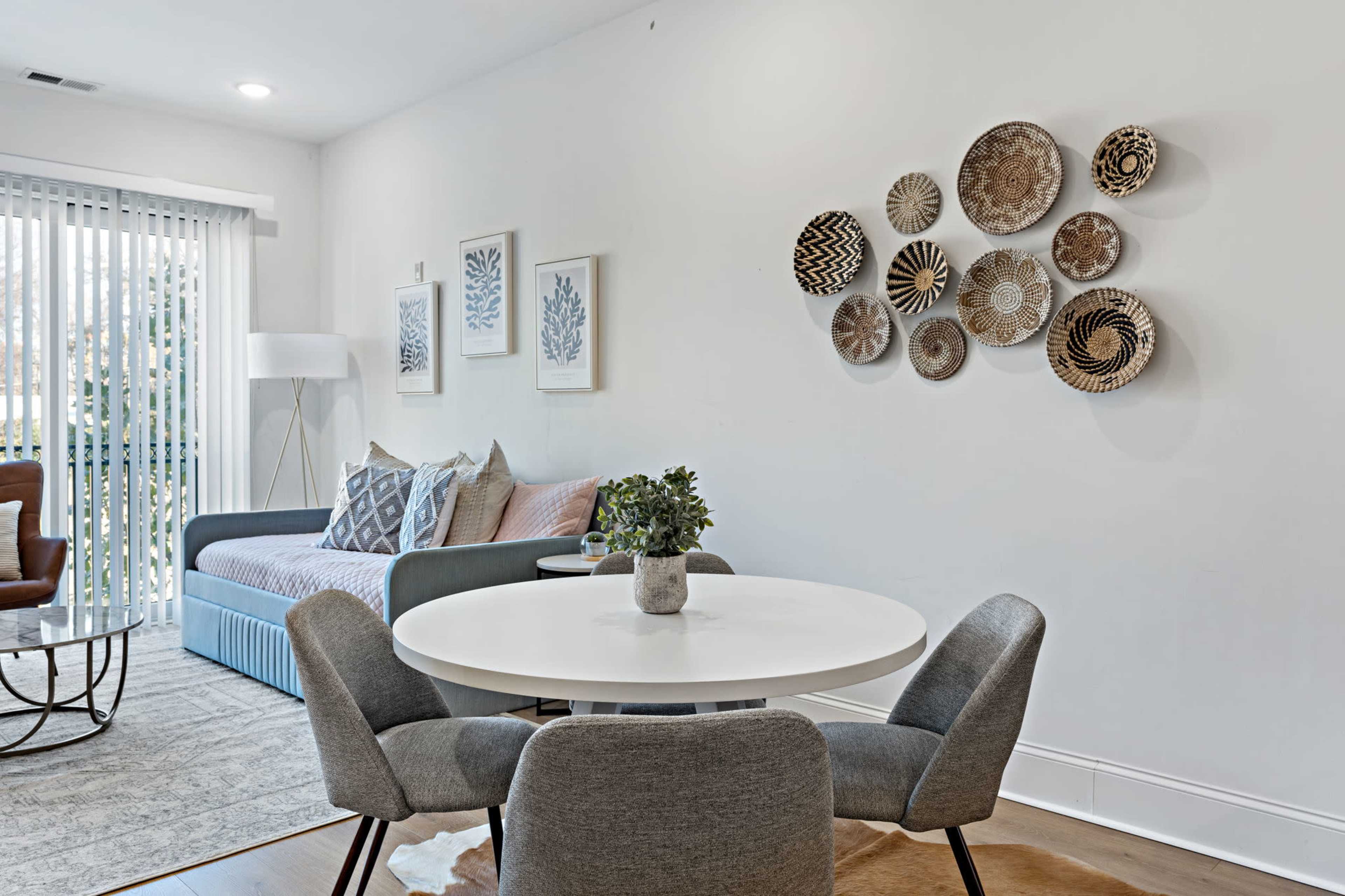 The image shows a modern living room featuring a sofa, decorative wall art made of woven plates, and a round dining table with gray chairs.