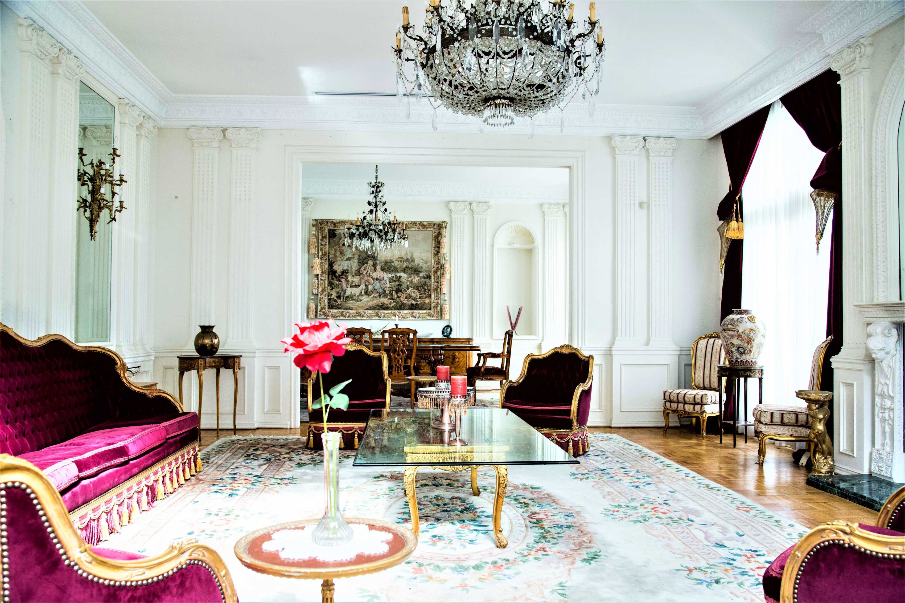 The room features ornate furniture, a crystal chandelier, and a large mirror, highlighted by a central glass table and a red rose in a vase.
