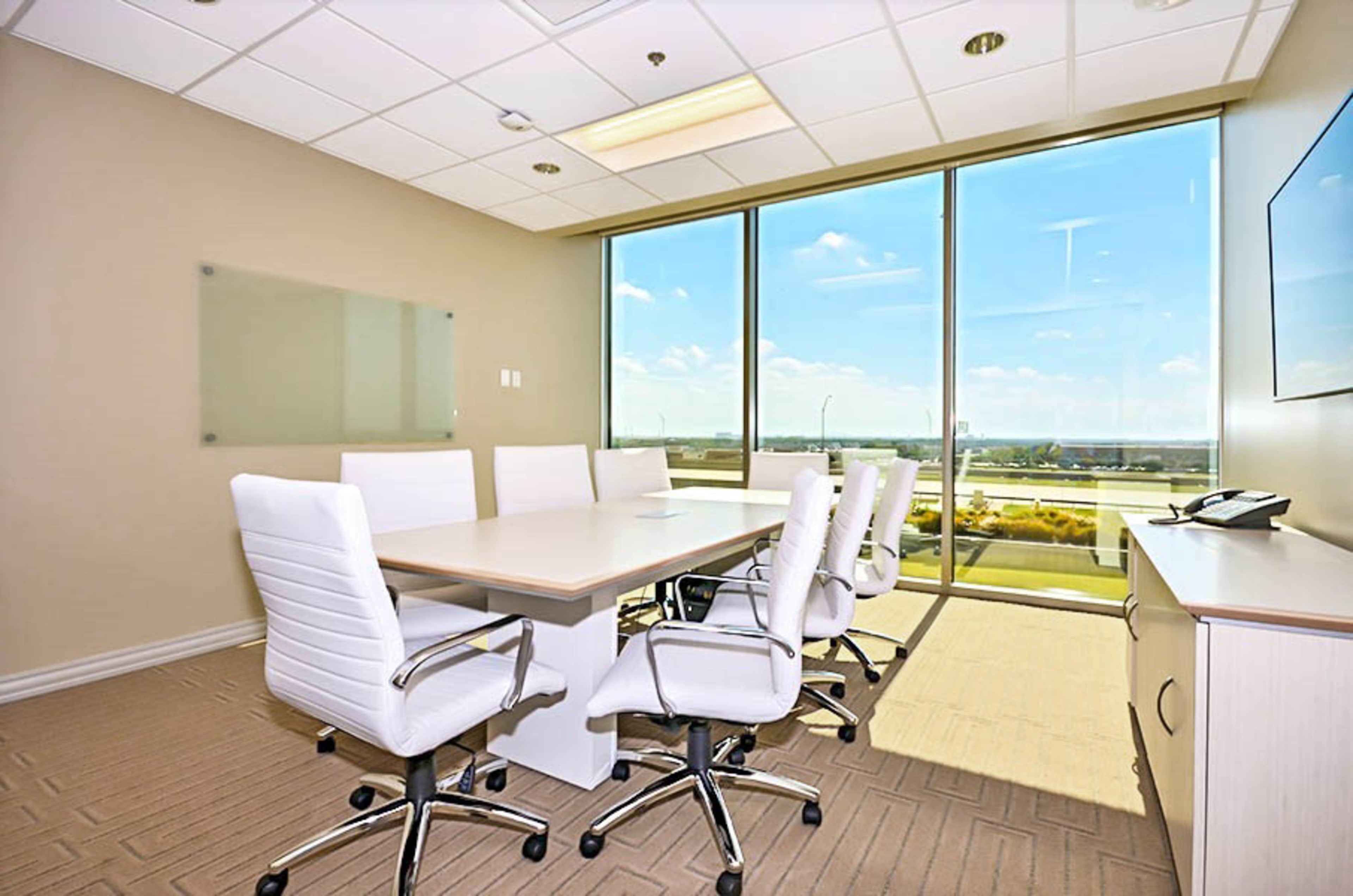 A modern conference room with a large table surrounded by white chairs, featuring a wall of windows that offer a view of the outdoors.