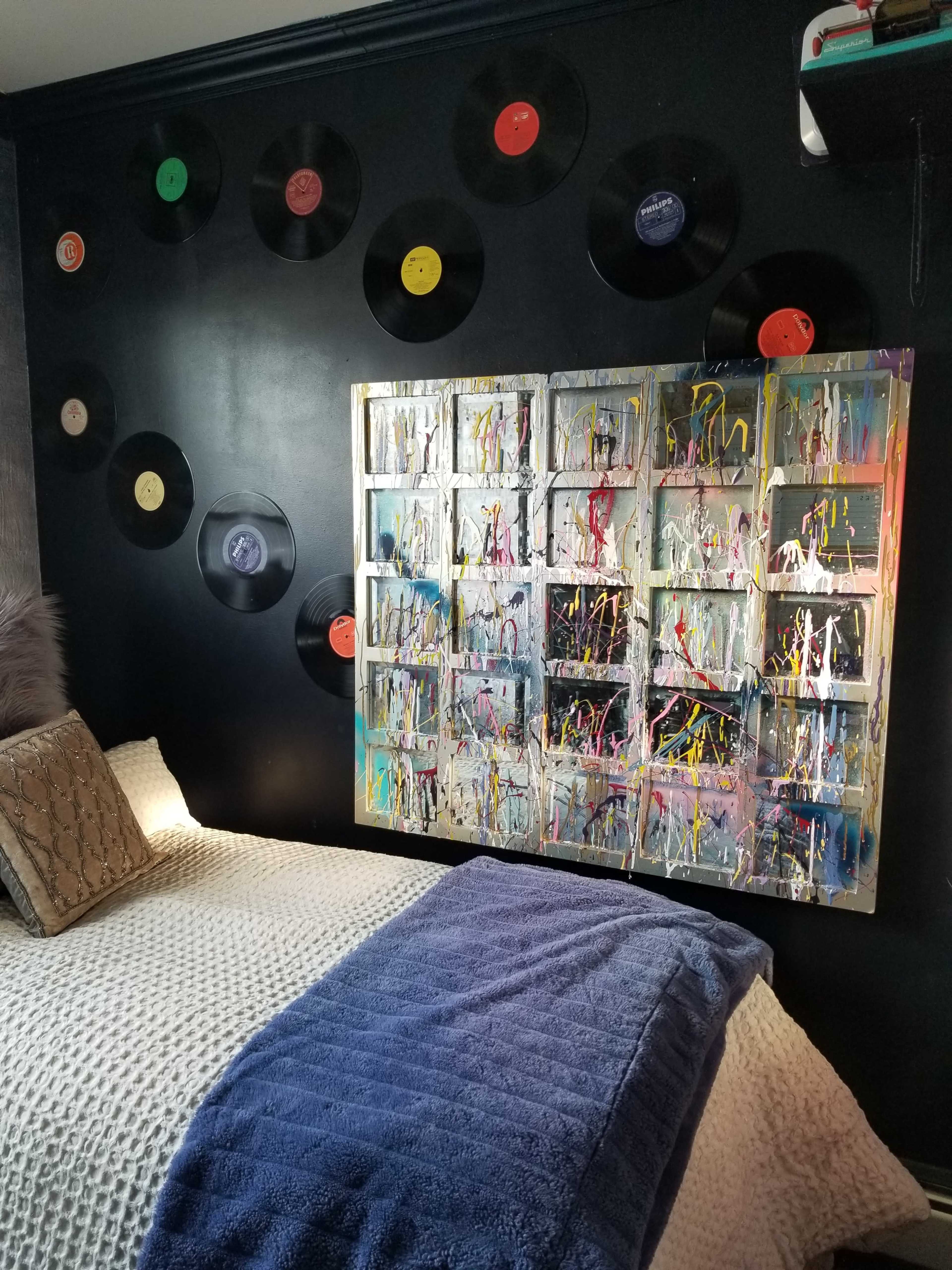 A bed with a textured white blanket is positioned against a dark wall adorned with colorful vinyl records and a large abstract painting.