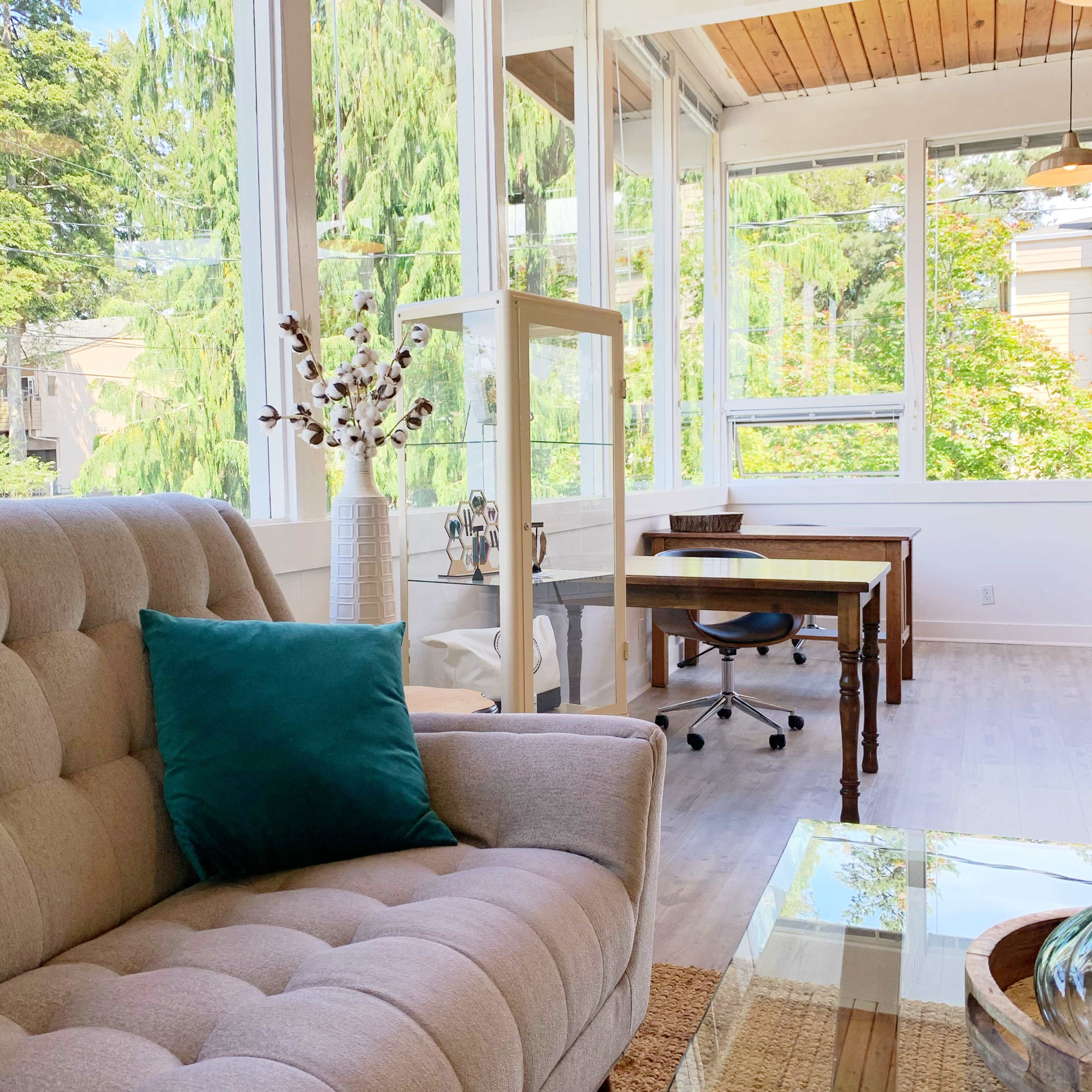 A bright sunroom with large windows, featuring a gray tufted sofa, a wooden desk with a chair, and decorative elements like a vase and potted plants.

