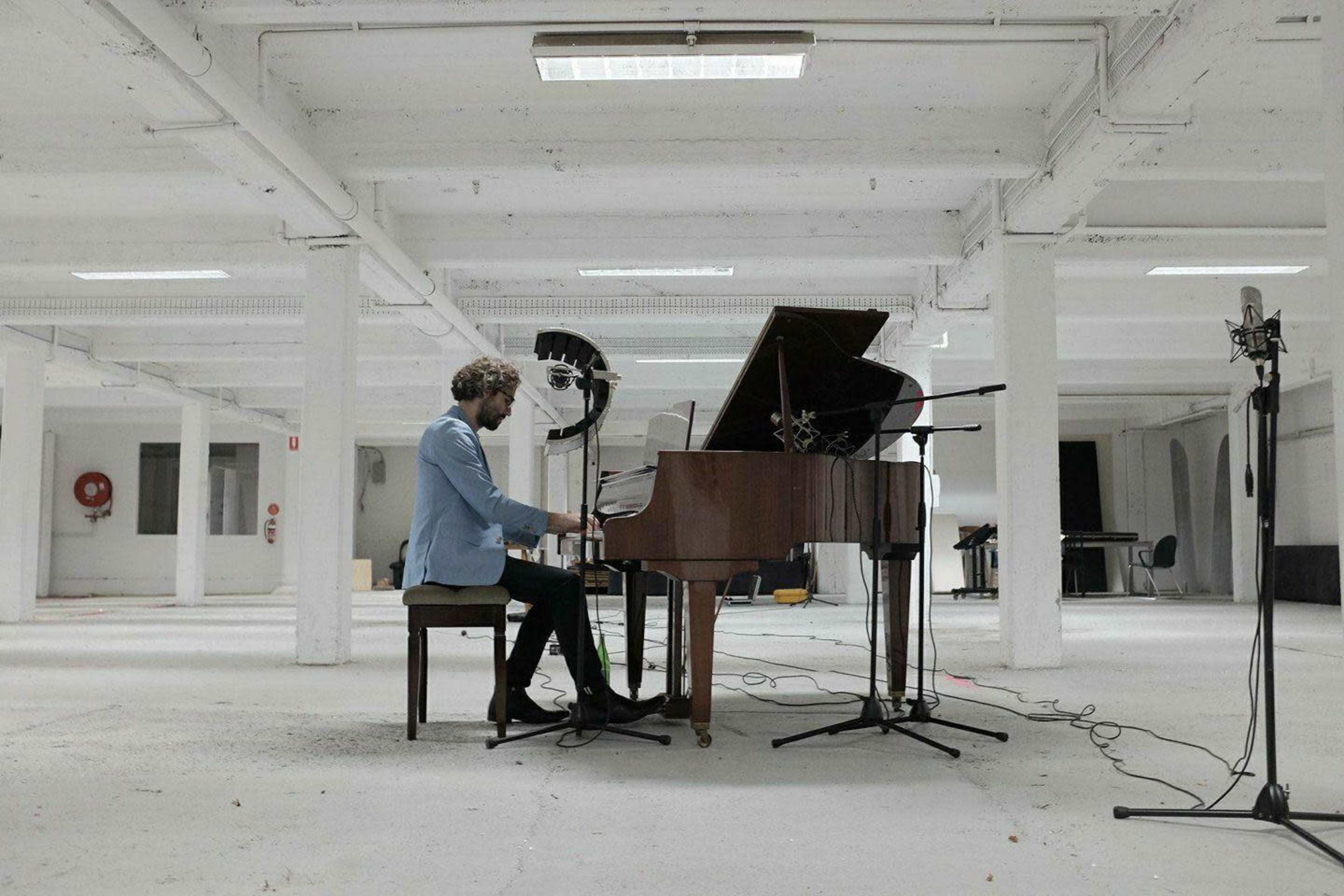 A musician plays a grand piano in a spacious, empty room with white walls and visible microphones set up around him.