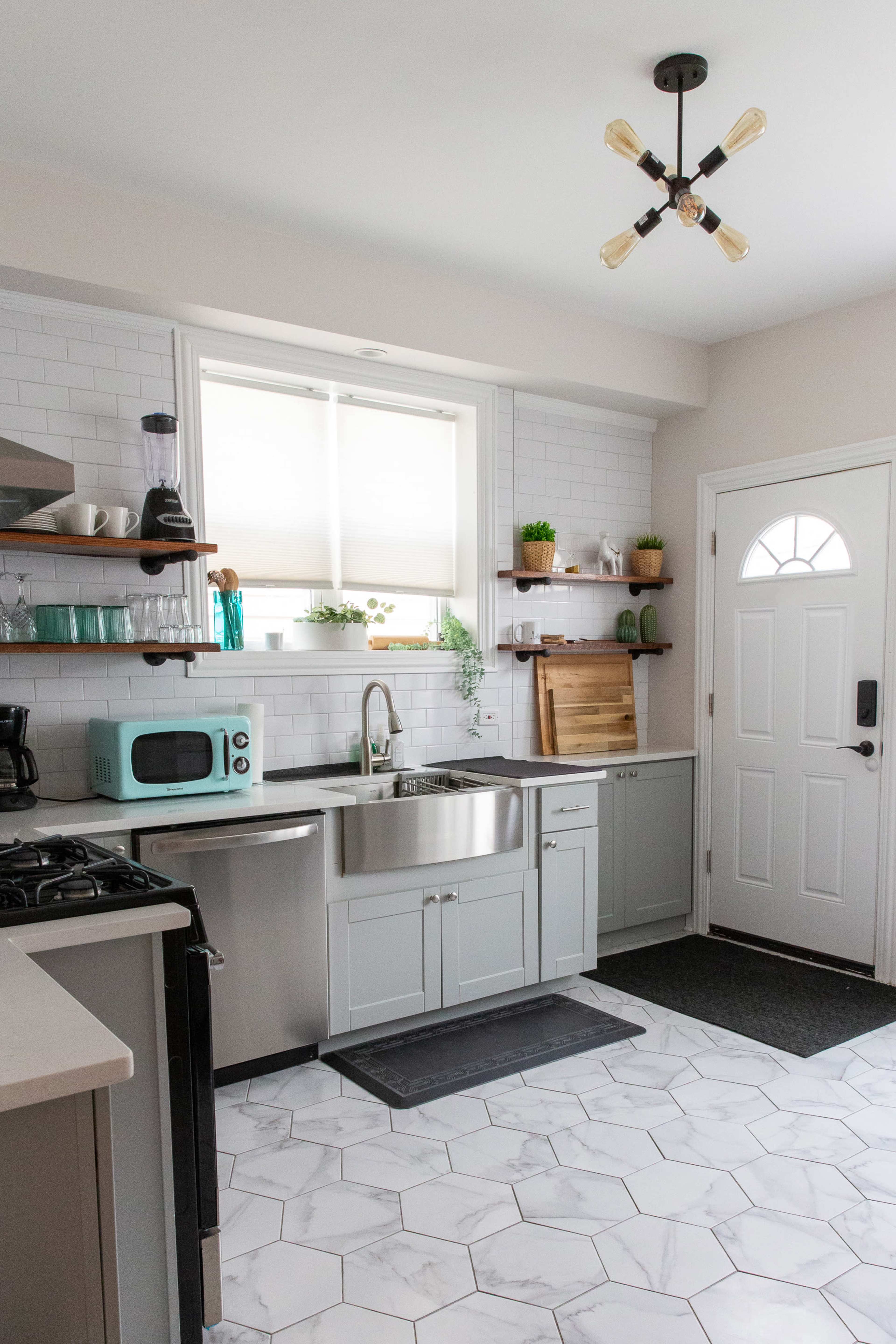 The kitchen features light gray cabinets, a stainless steel sink, a white tiled backsplash, and a door leading outside, with a window allowing natural light to enter.