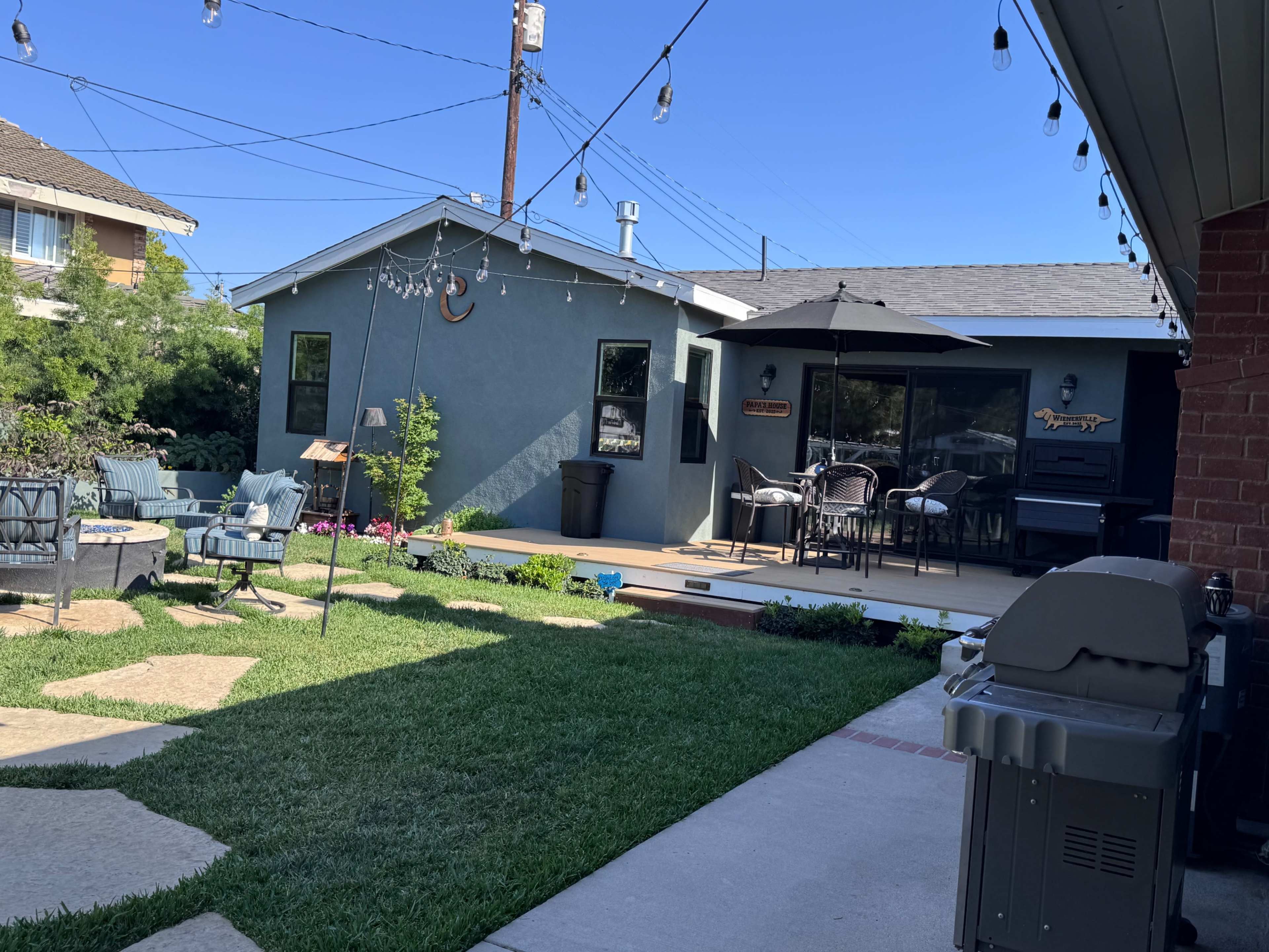 The image shows a well-maintained backyard featuring a patio with outdoor furniture, a barbecue grill, and a blue house alongside a garden area with decorative lights.