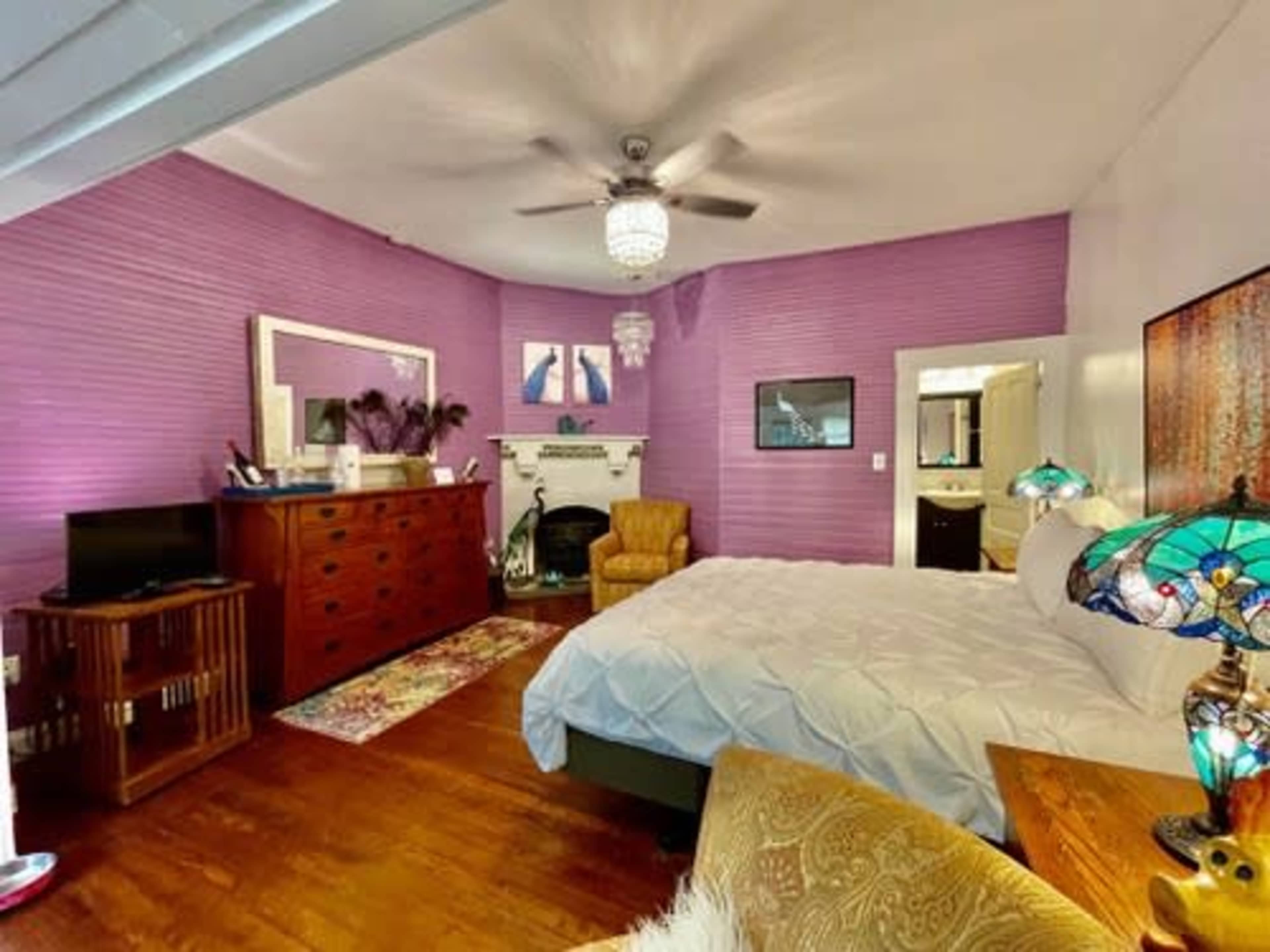 A bedroom features a purple-paneled wall, a large bed, a dresser, a vintage armchair, and stained glass lamps.