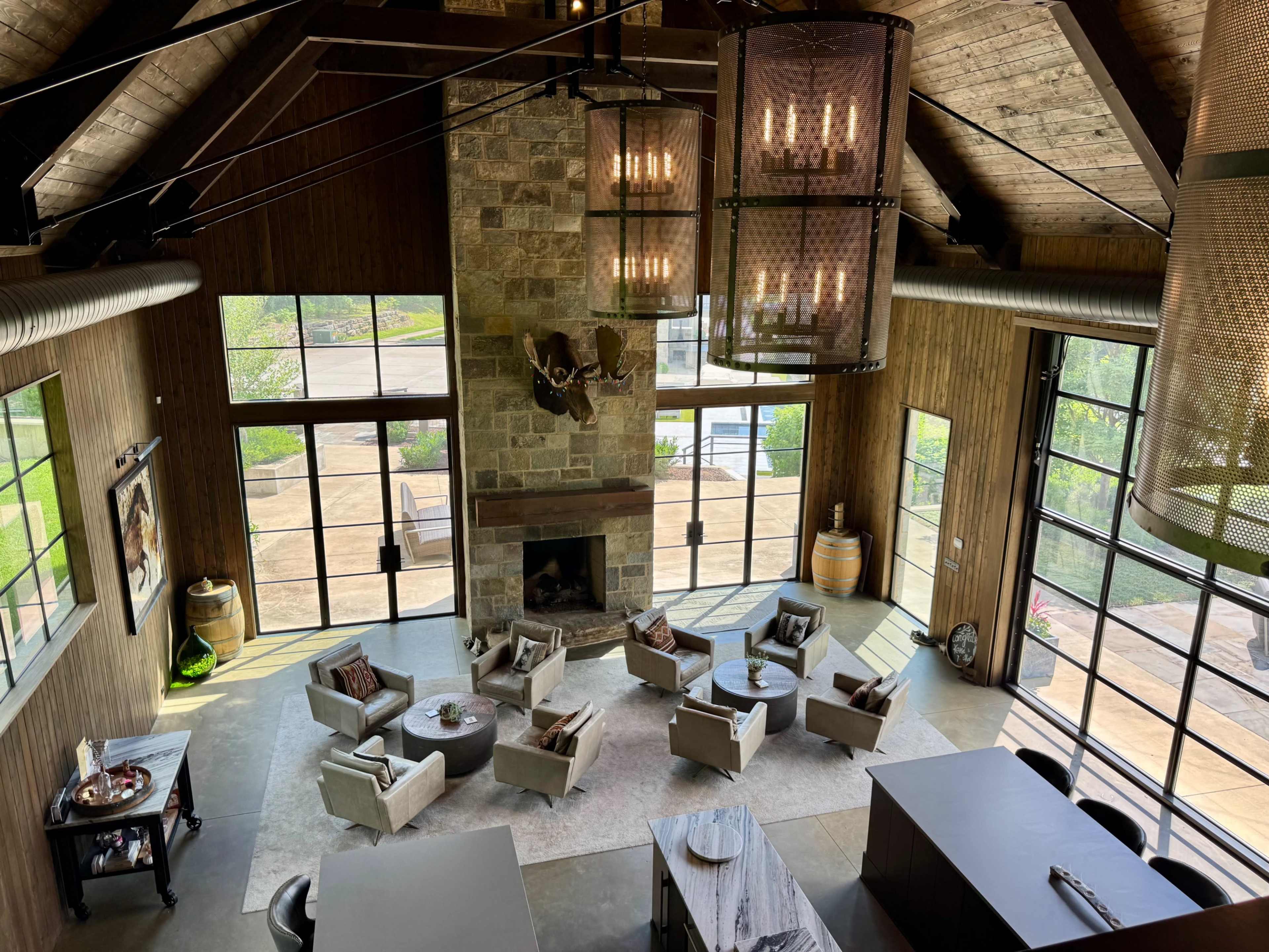 The image shows a spacious, modern living area with a high ceiling, large windows, and a stone fireplace, arranged with comfortable seating and a coffee table.