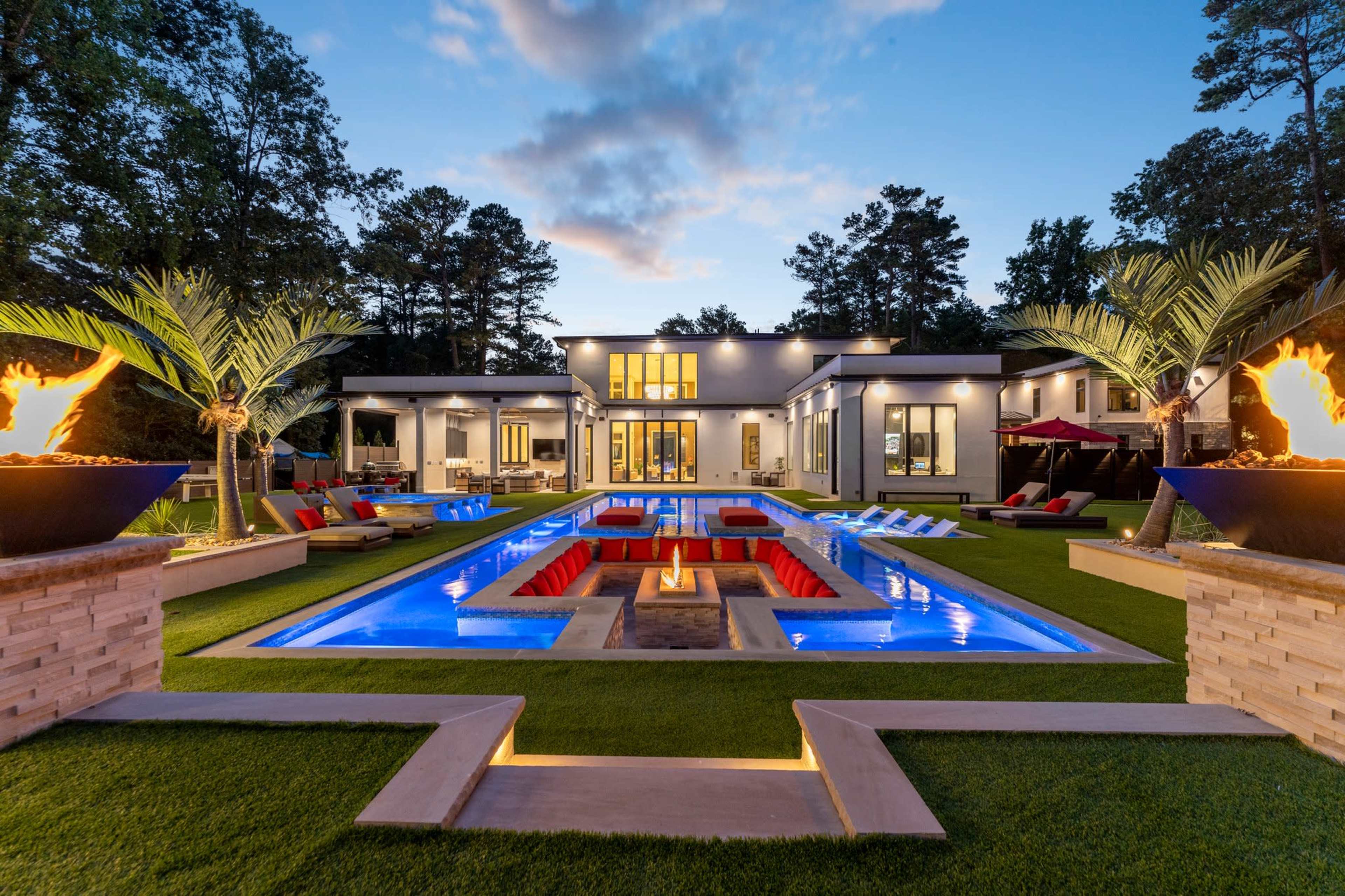 The image shows a modern house with a swimming pool surrounded by palm trees and illuminated fire features, set in a landscaped yard during twilight.