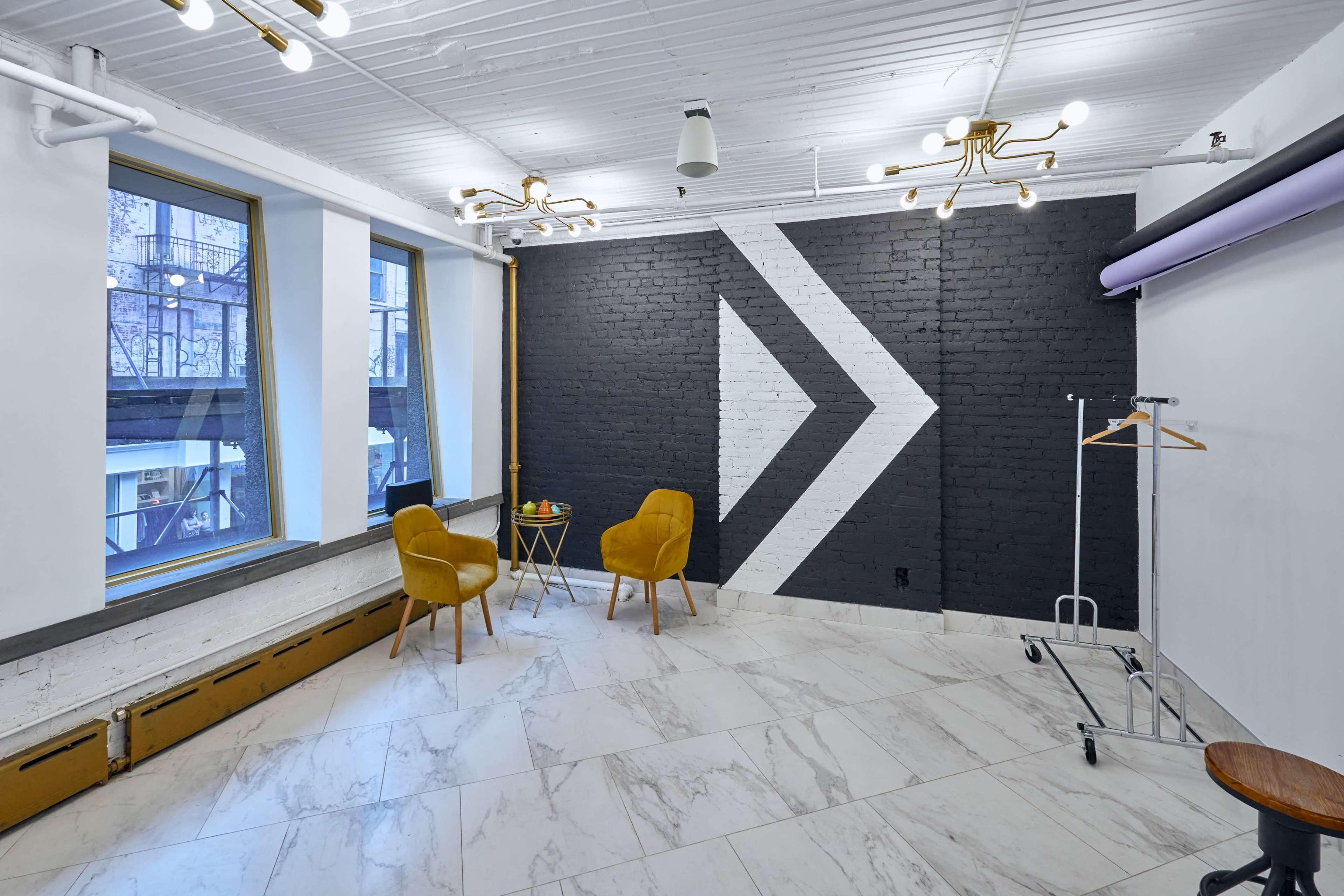 A modern interior space featuring a black and white accent wall with an arrow design, two yellow chairs, a small table with fruit, and large windows.