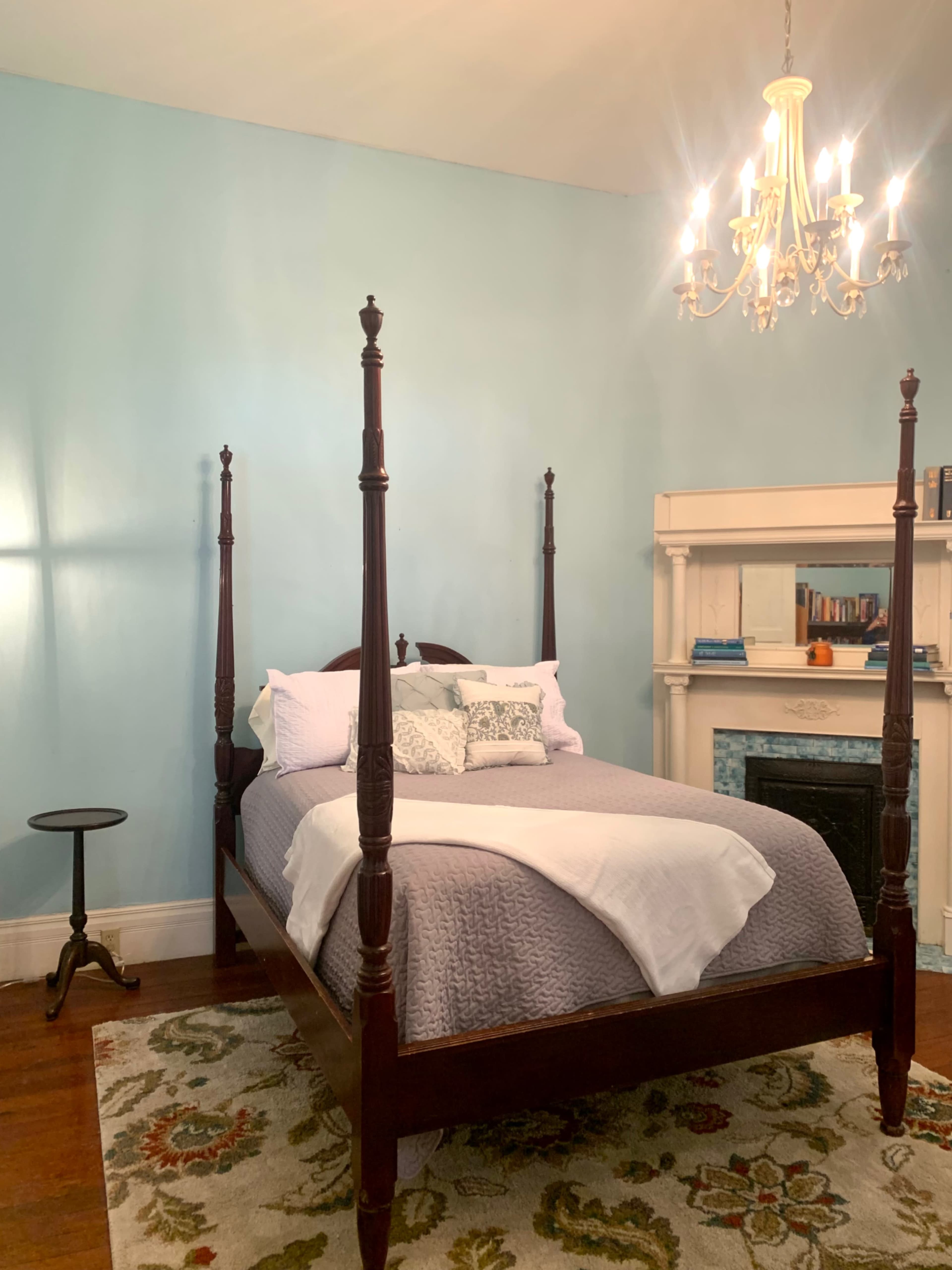The image shows a room with a four-poster bed, a small round table, and a decorative area rug, all under a chandelier.