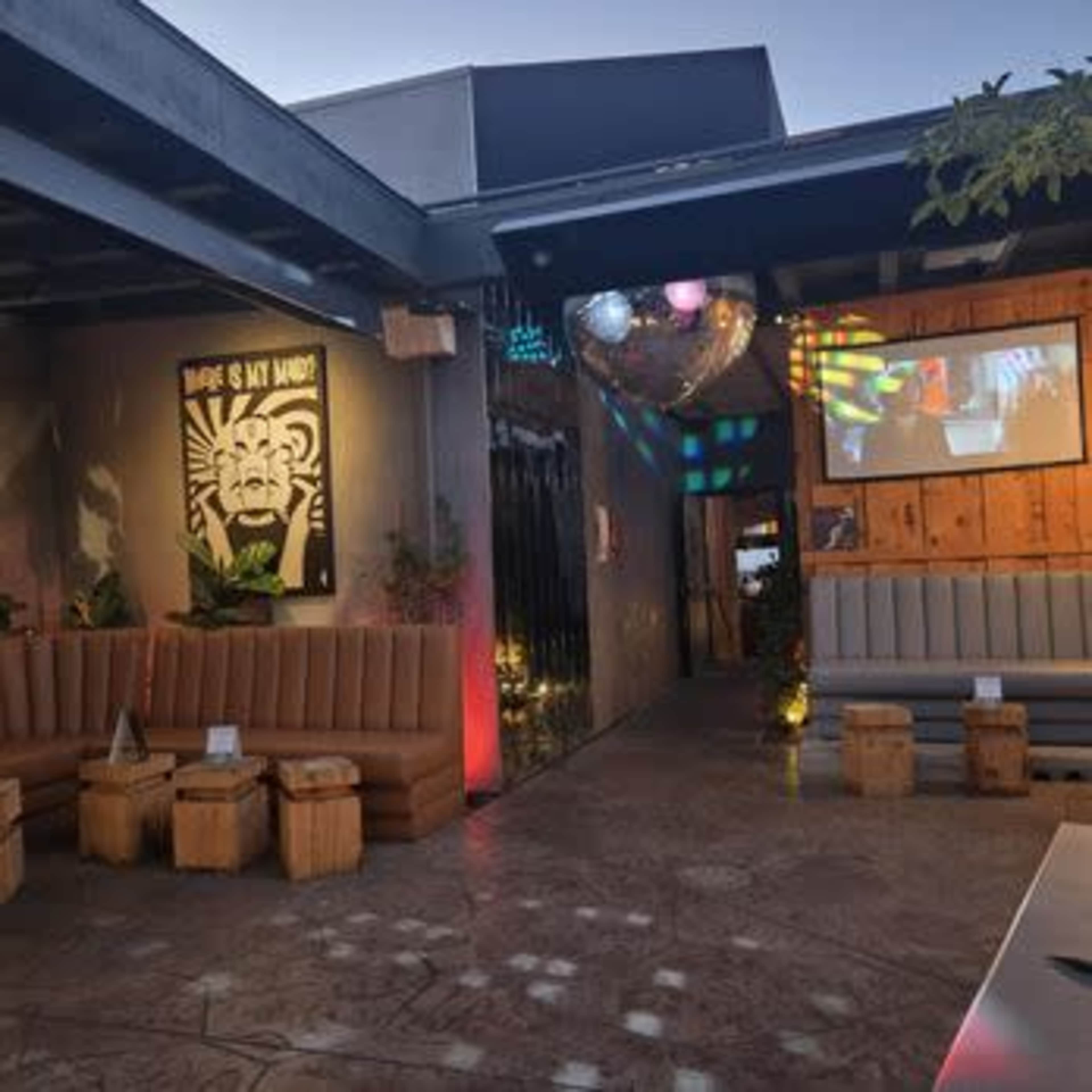 The image shows a lively outdoor bar area with seating, decorative lighting, and a large screen displaying images on a wall.