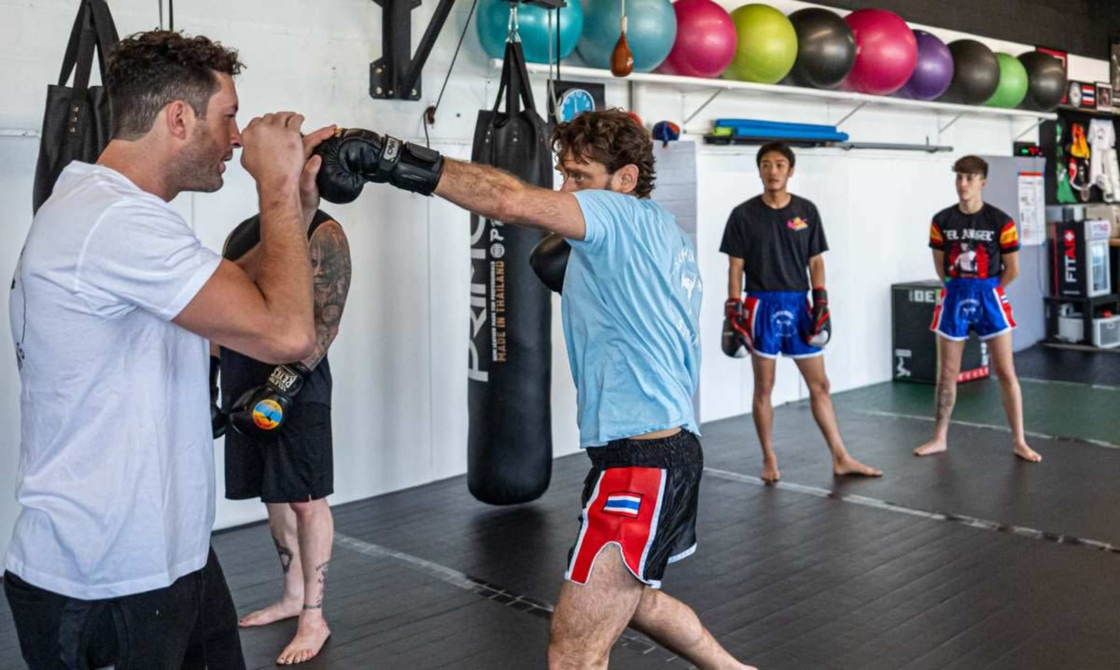 Two men are practicing striking techniques while two others observe in a gym setting equipped with punching bags and exercise balls.