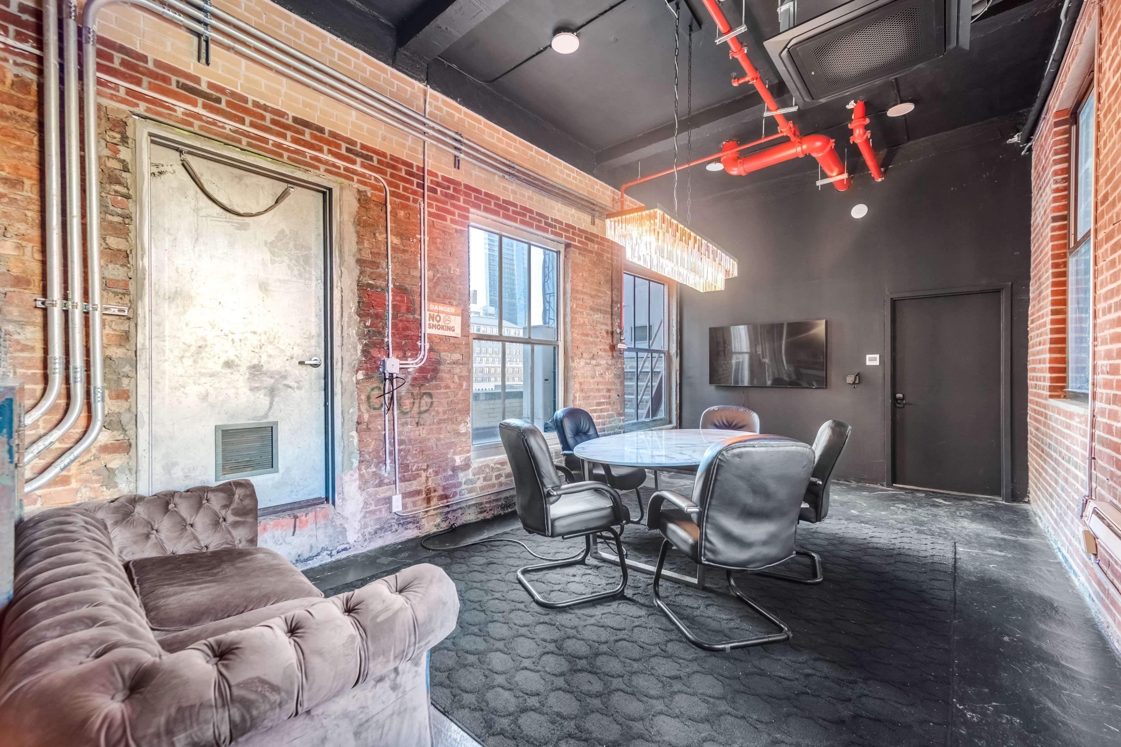 A modern conference room with brick walls, a round table surrounded by black leather chairs, and a large window offering a city view.