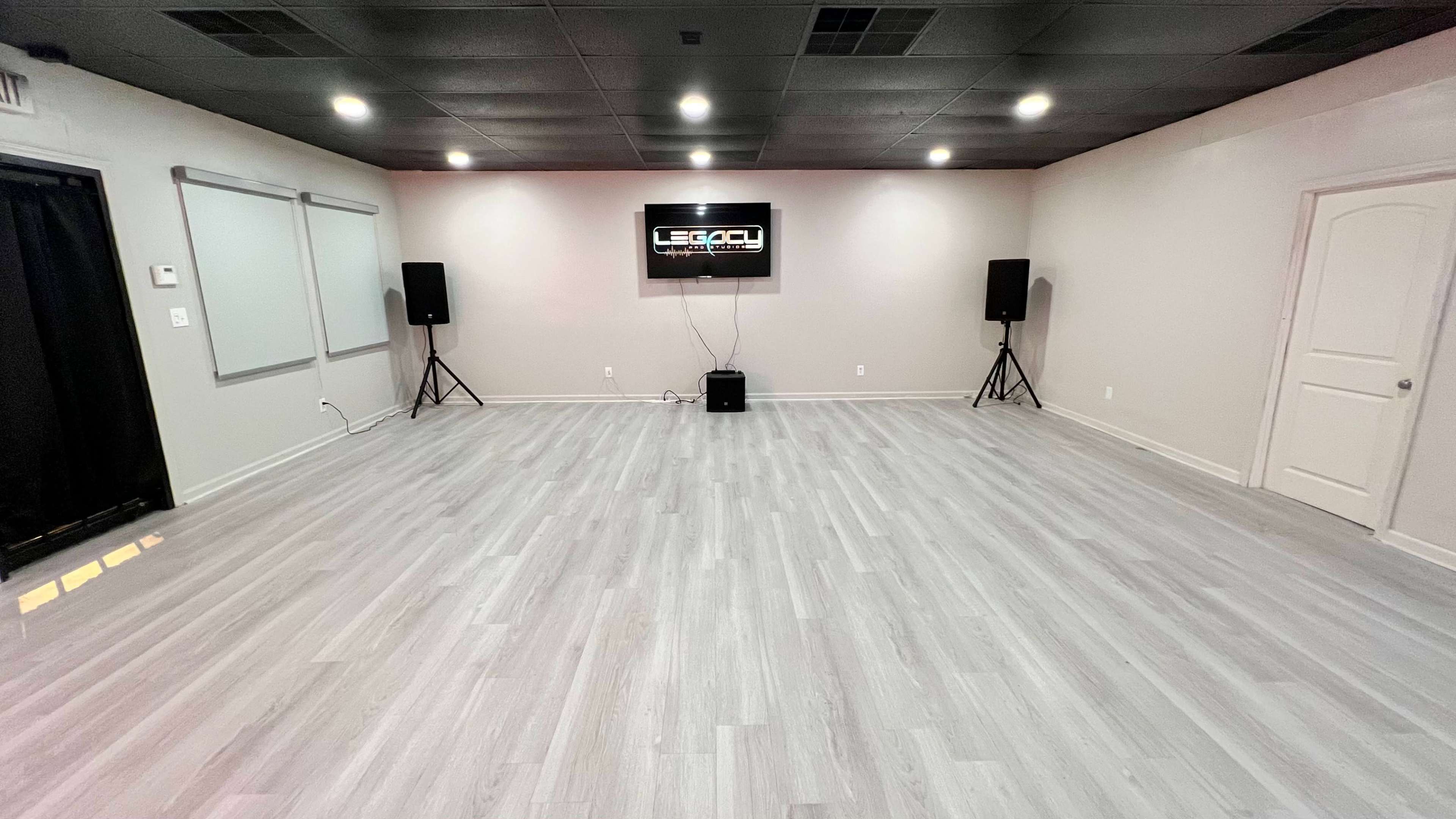The image shows a spacious, empty room with pale wooden flooring, two speakers on stands, and a wall-mounted screen displaying the word "LEGACY."