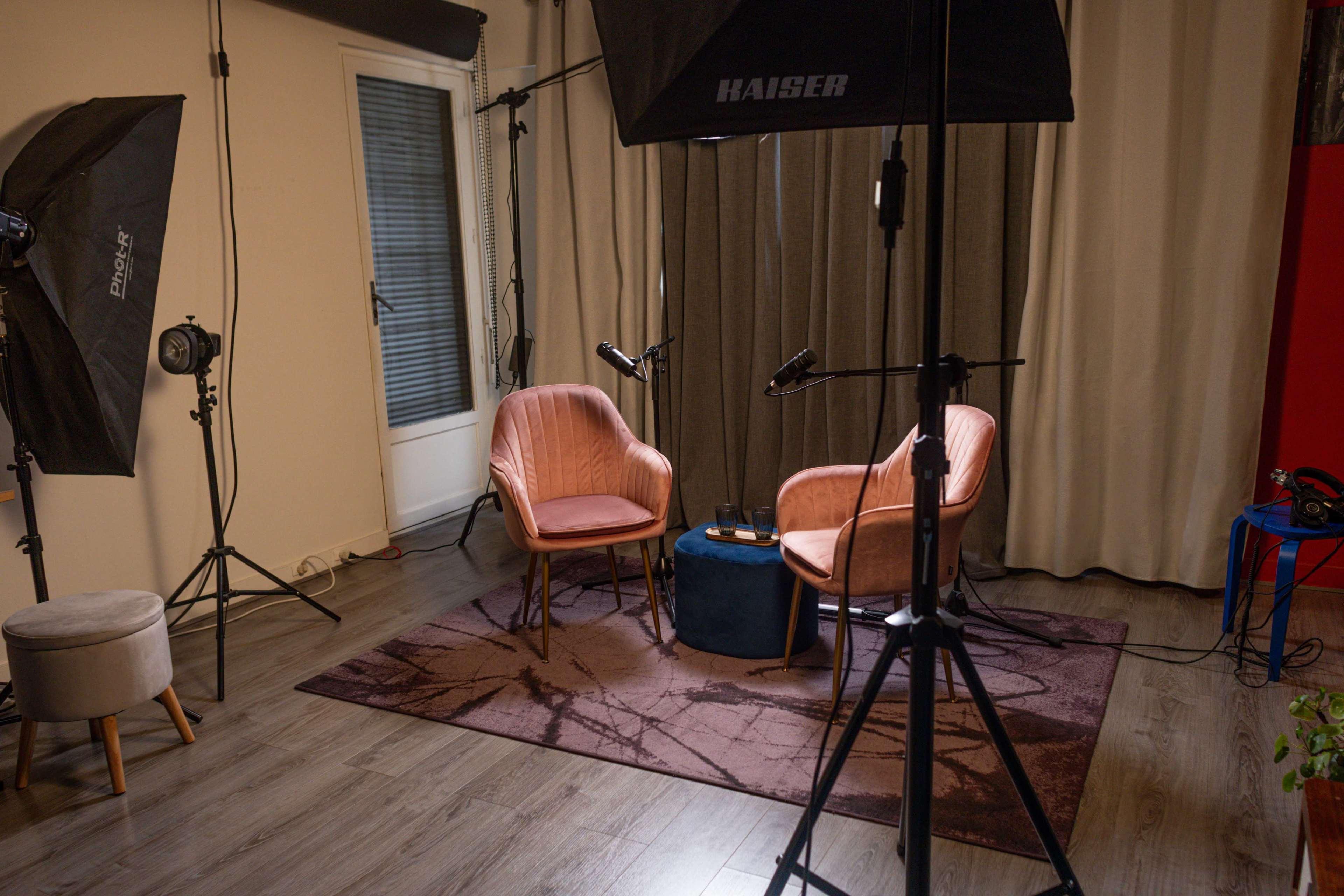 The image shows a small studio setup featuring two pink chairs facing each other on a patterned rug, with professional lighting equipment positioned around the area.