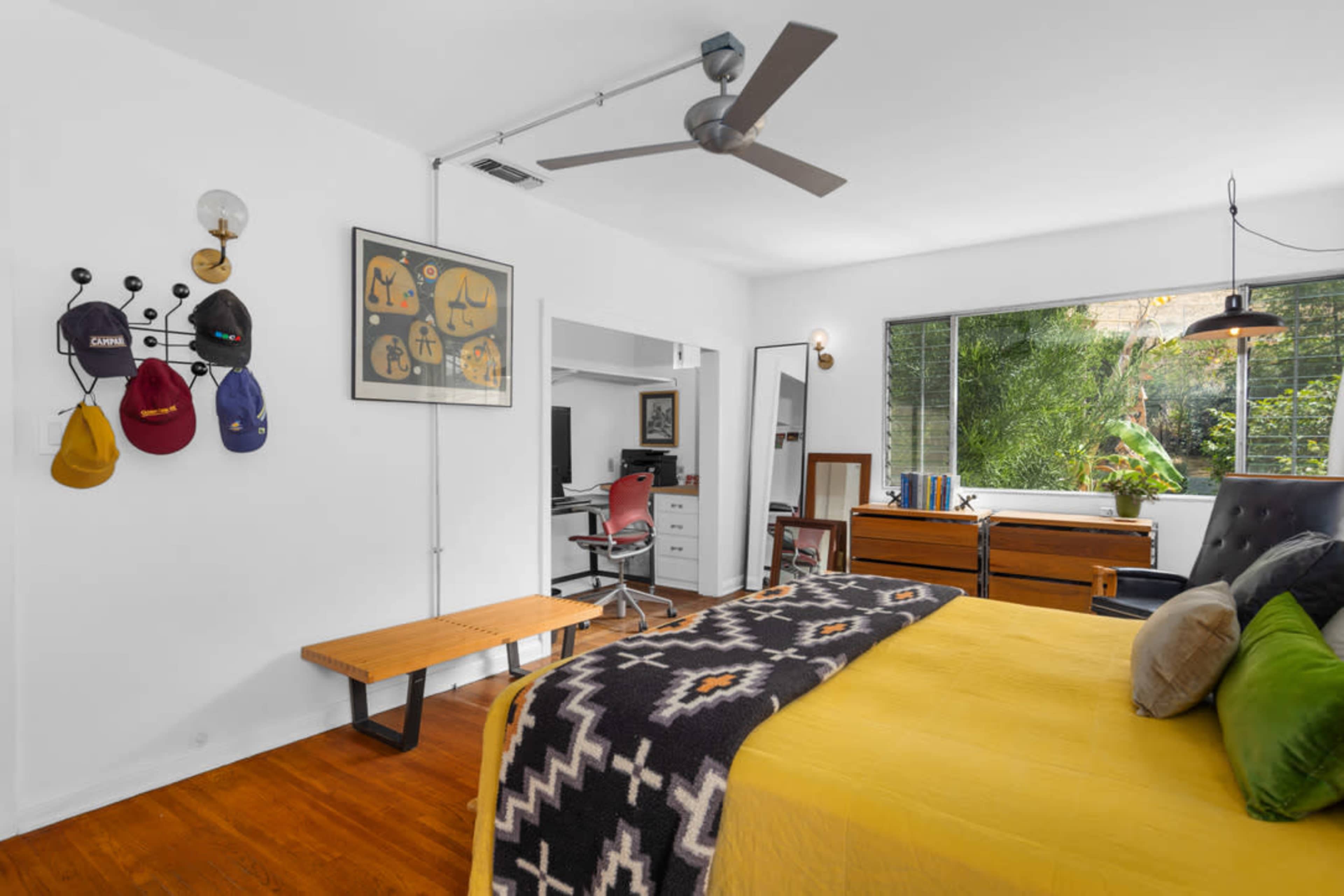 The image shows a modern bedroom with a yellow bedspread, a wall display of hats, a desk with a computer, and large windows overlooking greenery.