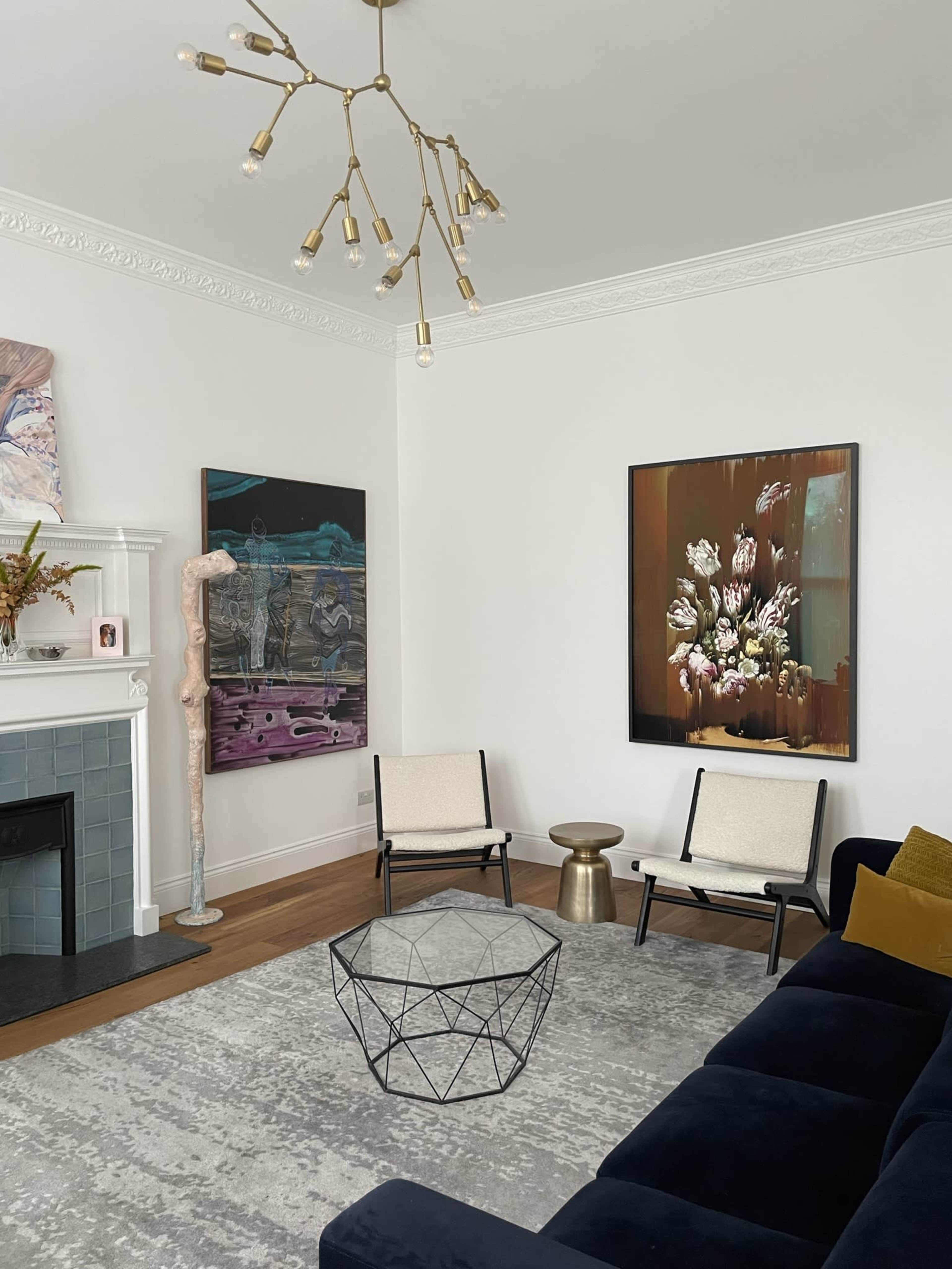 The image shows a modern living room with two chairs and a coffee table, featuring a fireplace, two large artworks on the walls, and a unique light fixture.