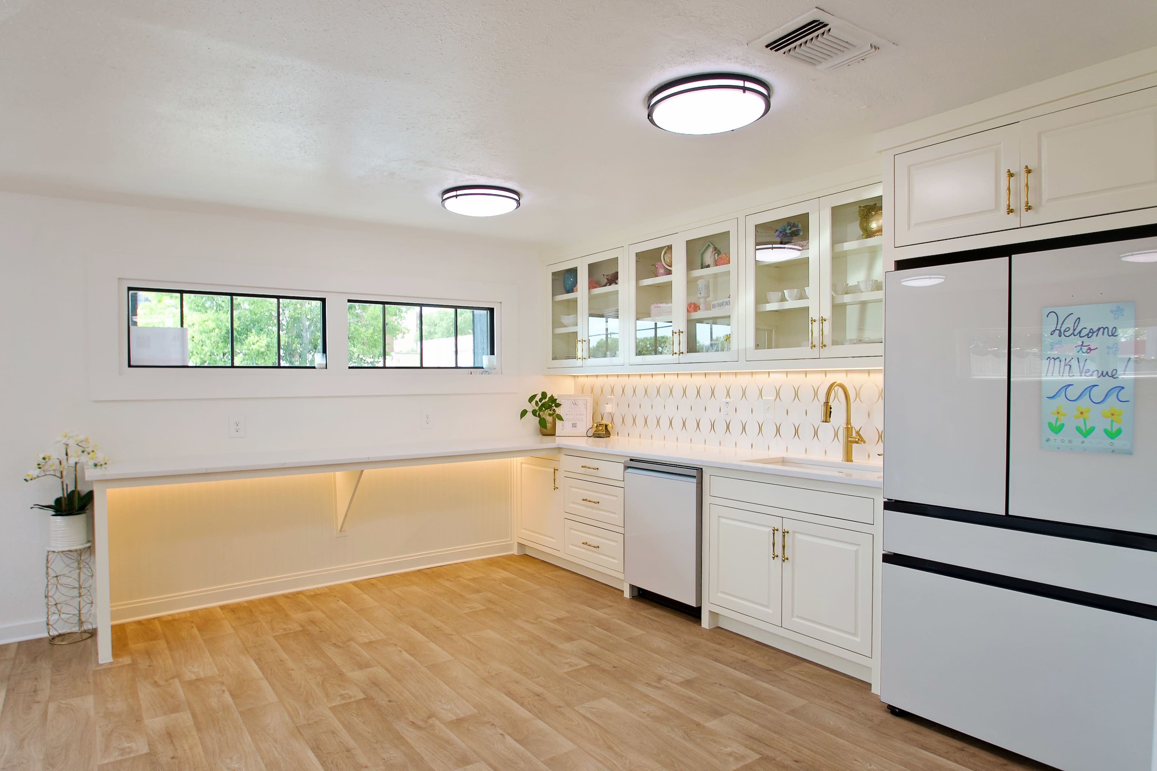 The image shows a modern kitchen featuring white cabinetry, a stainless steel refrigerator, and a countertop with under-cabinet lighting.