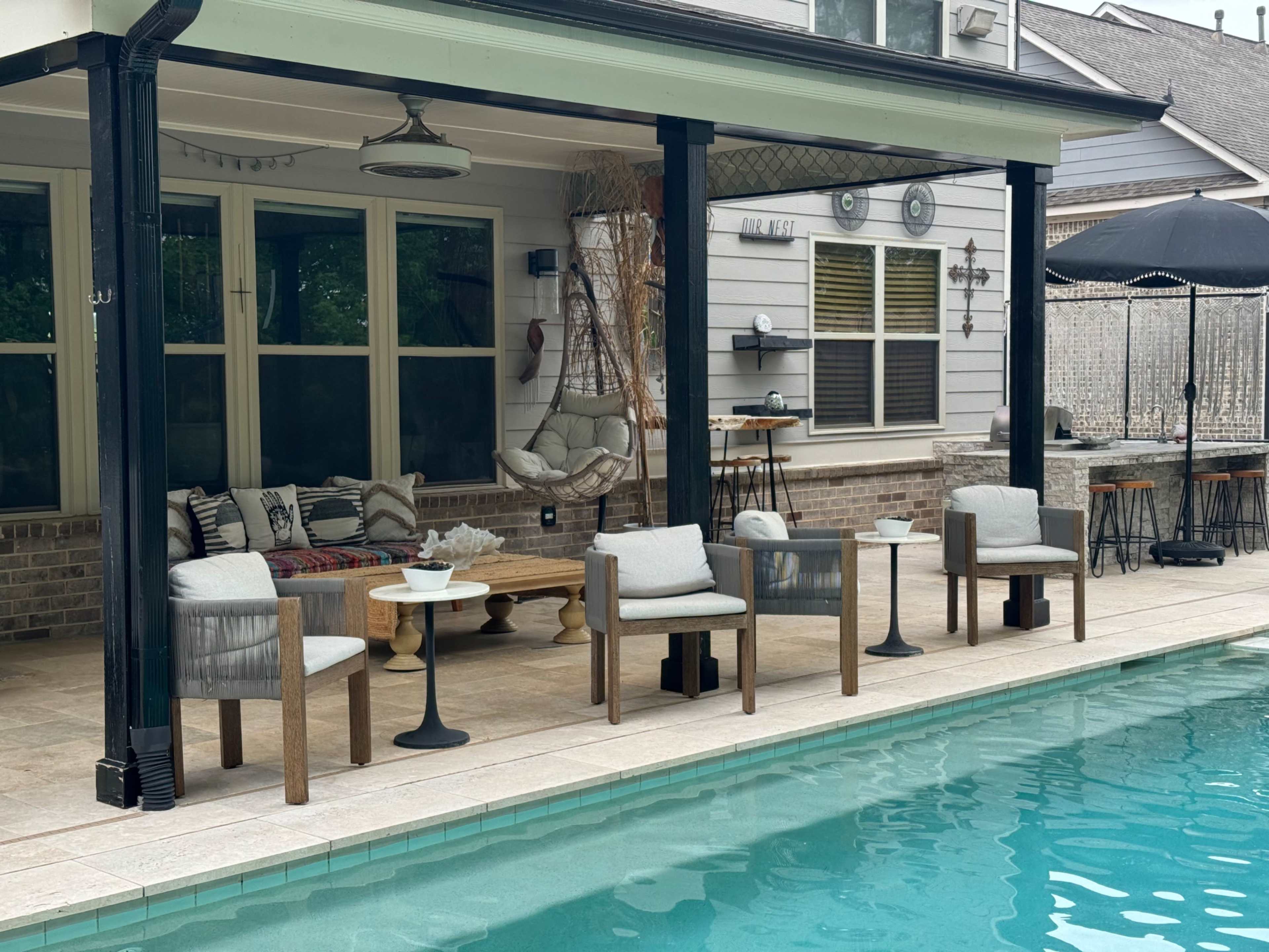 A poolside patio features several modern chairs and small tables arranged next to a swimming pool, with a covered seating area in the background.