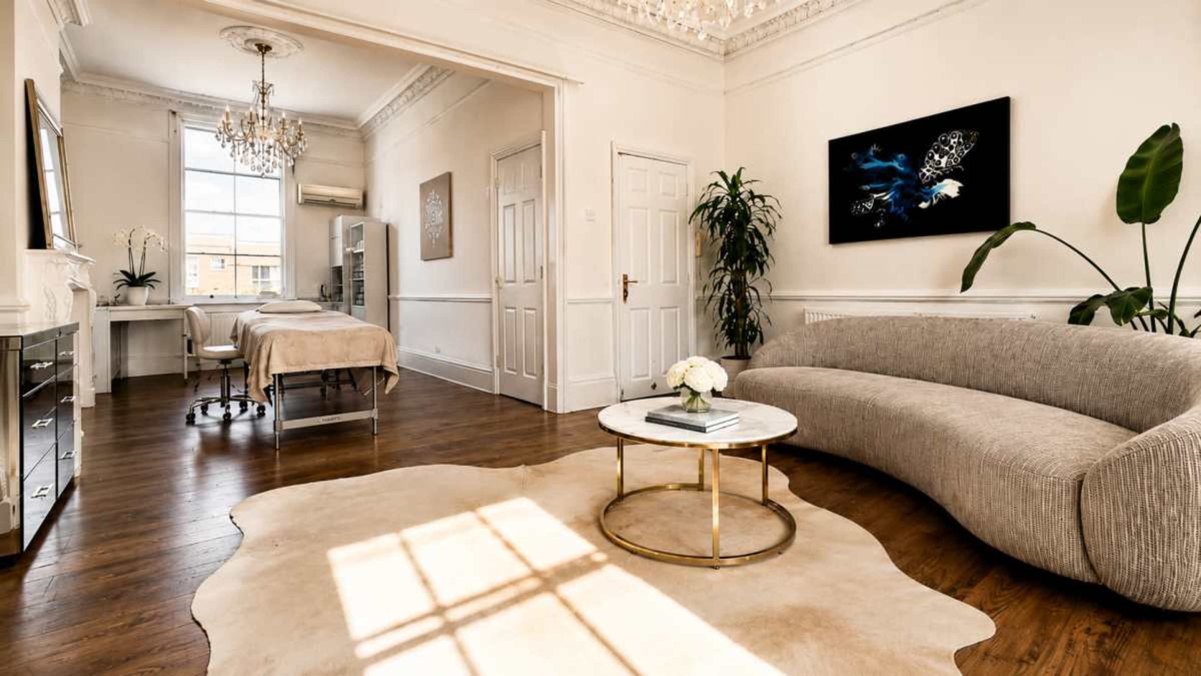 The image shows a spacious, elegantly decorated room with a curved gray sofa, a round coffee table, and a treatment bed in a well-lit environment featuring natural light and a chandelier.