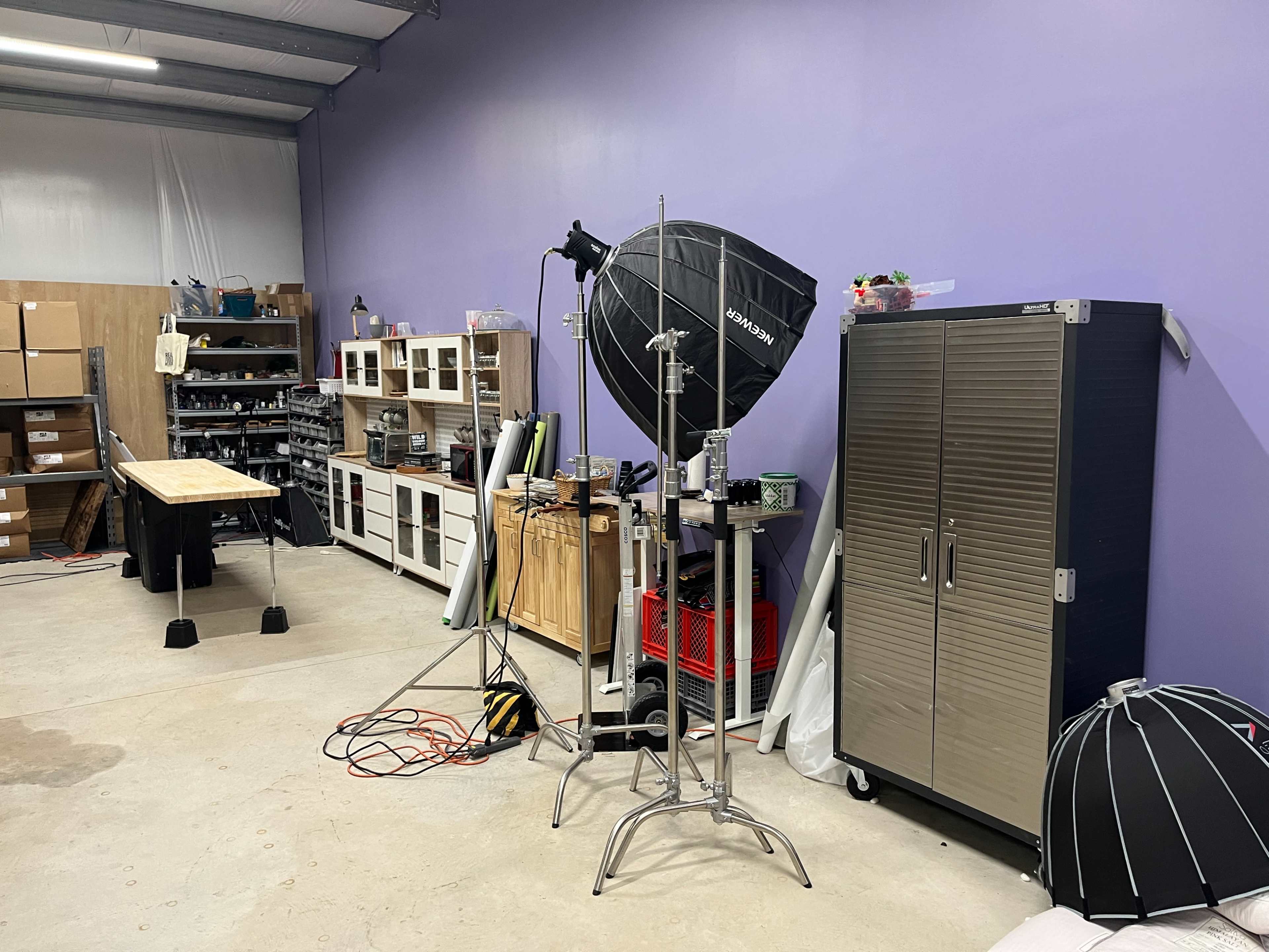 A photography studio with various equipment, including a large softbox, a table, storage units, and a cabinet along a purple wall.