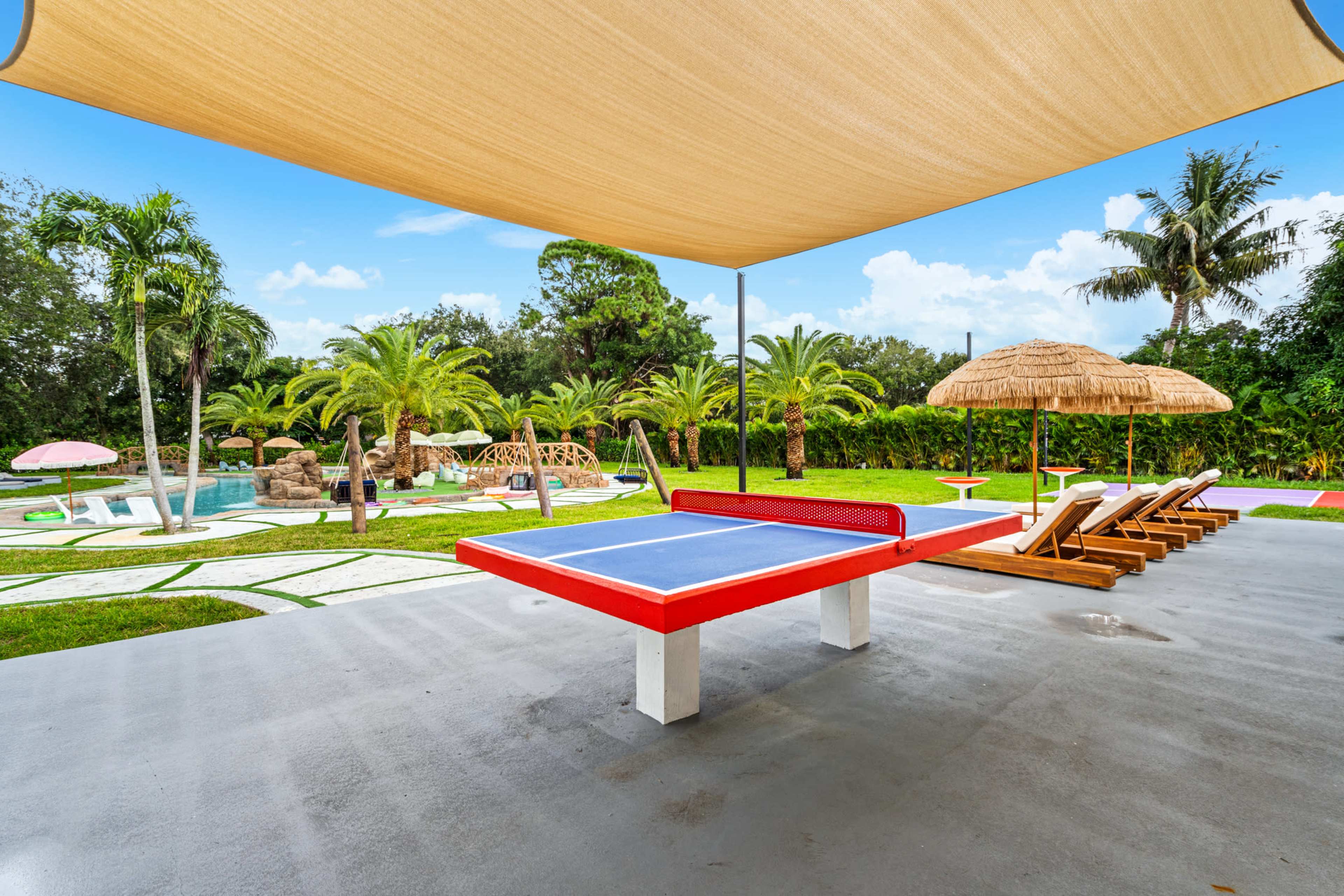 A blue ping pong table is set under a shade tarp beside a swimming pool, surrounded by palm trees and lounge chairs.