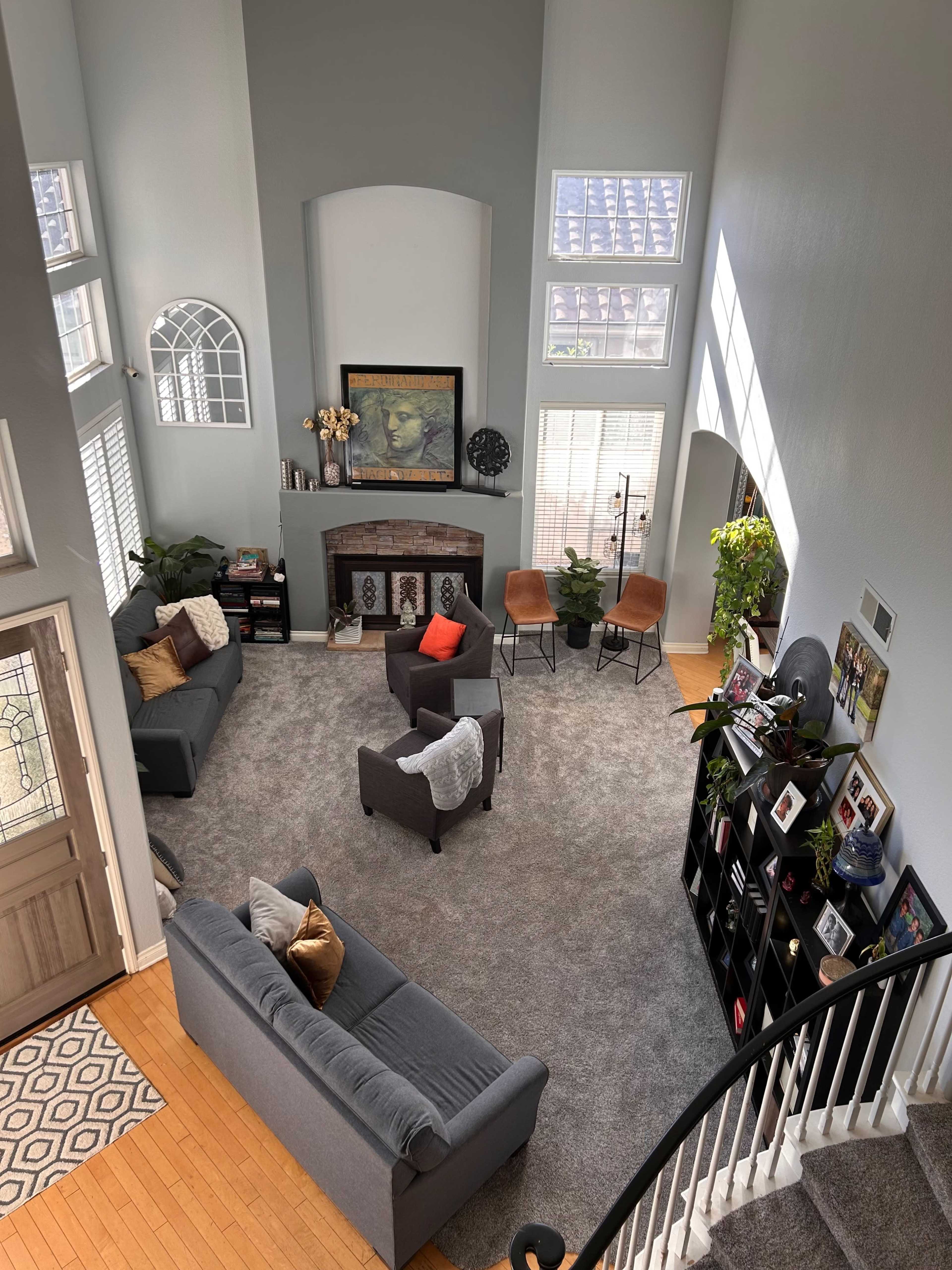 A spacious living room with high ceilings, multiple windows, a fireplace, and seating arranged around a central area on a gray carpet.