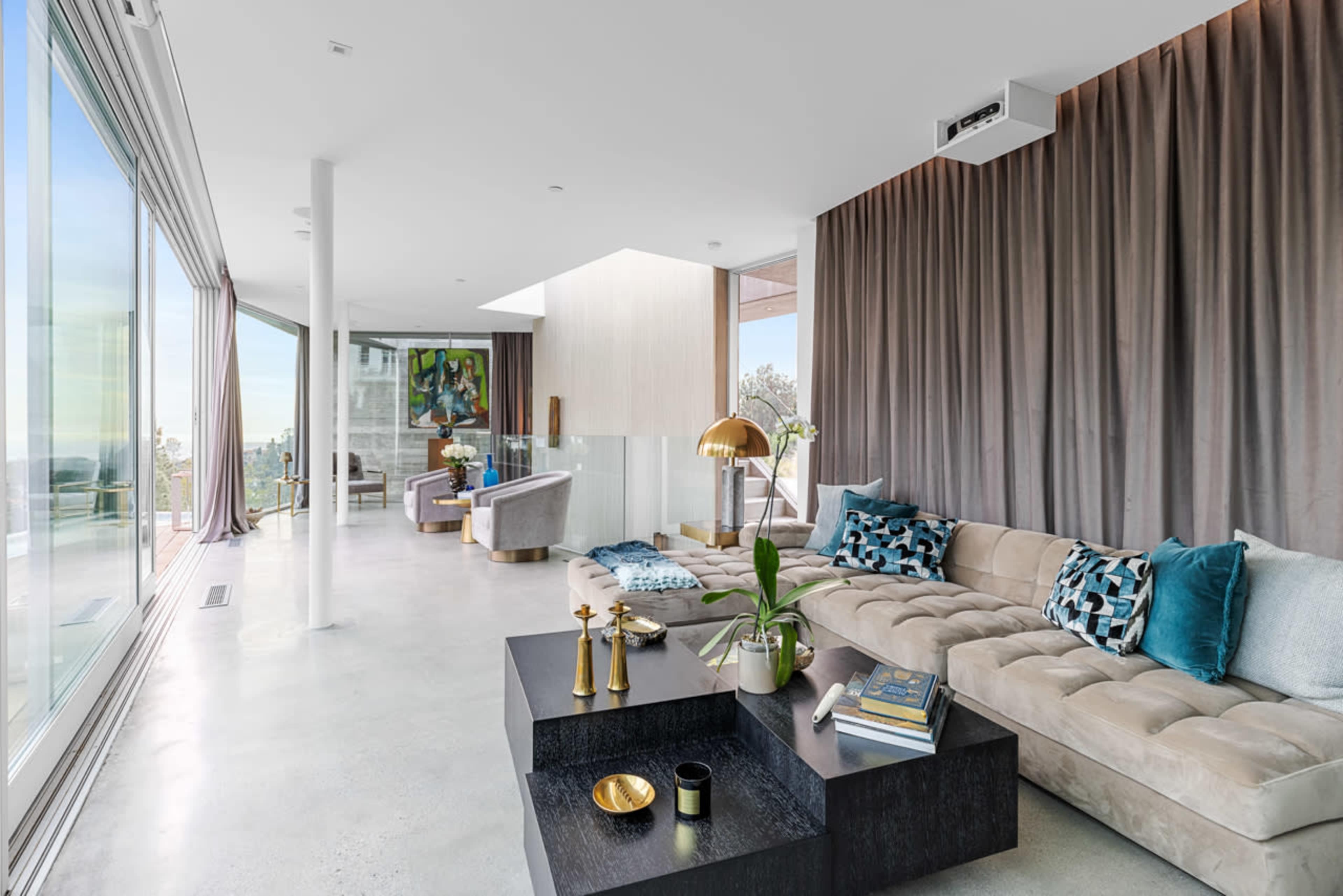 The image shows a modern living room with a large sectional sofa, eclectic decor, and expansive windows that offer a view of the outdoors.