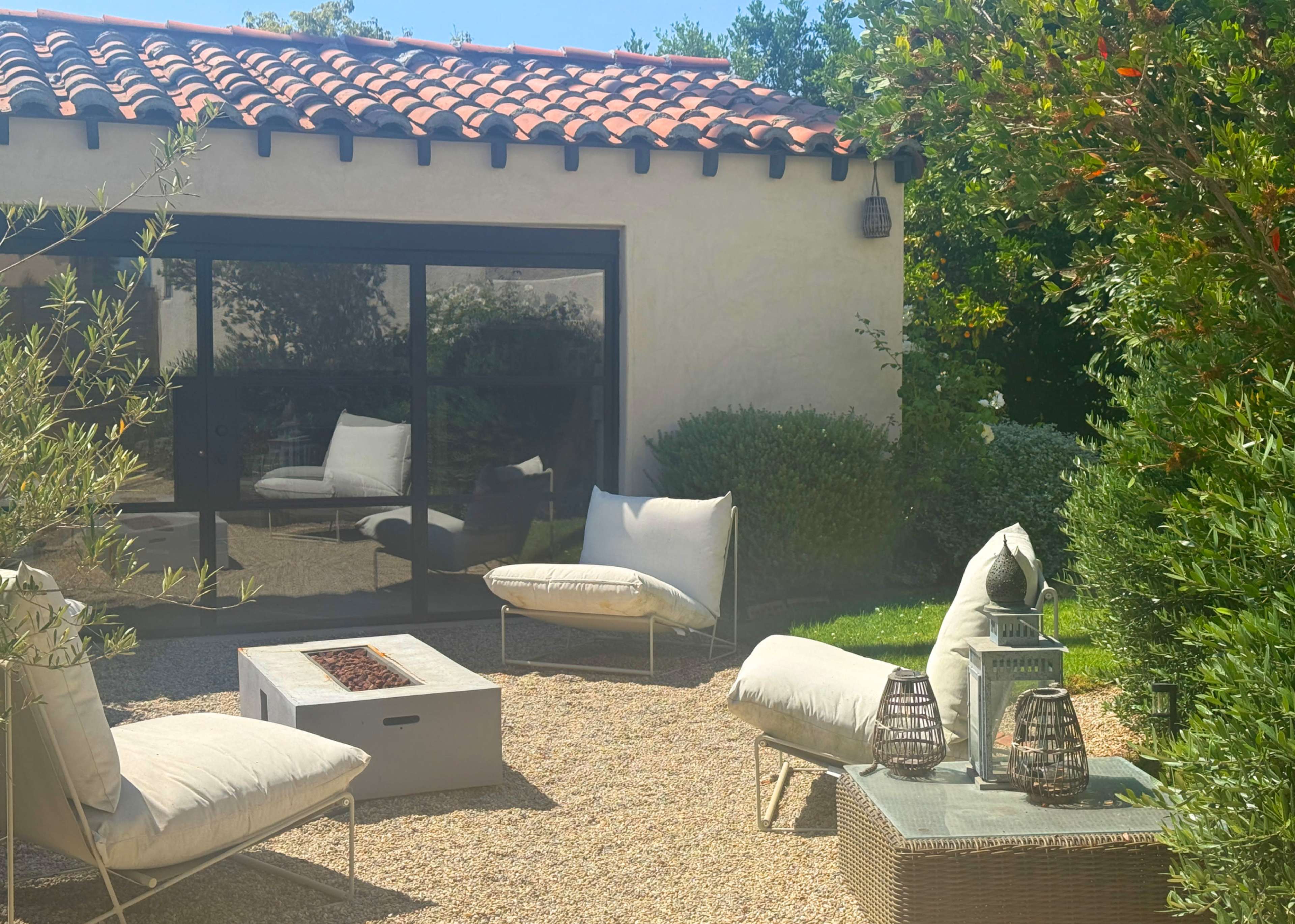 A sunlit outdoor area features a modern gravel patio with several cushioned seating options, a fire pit, and surrounded by lush greenery.