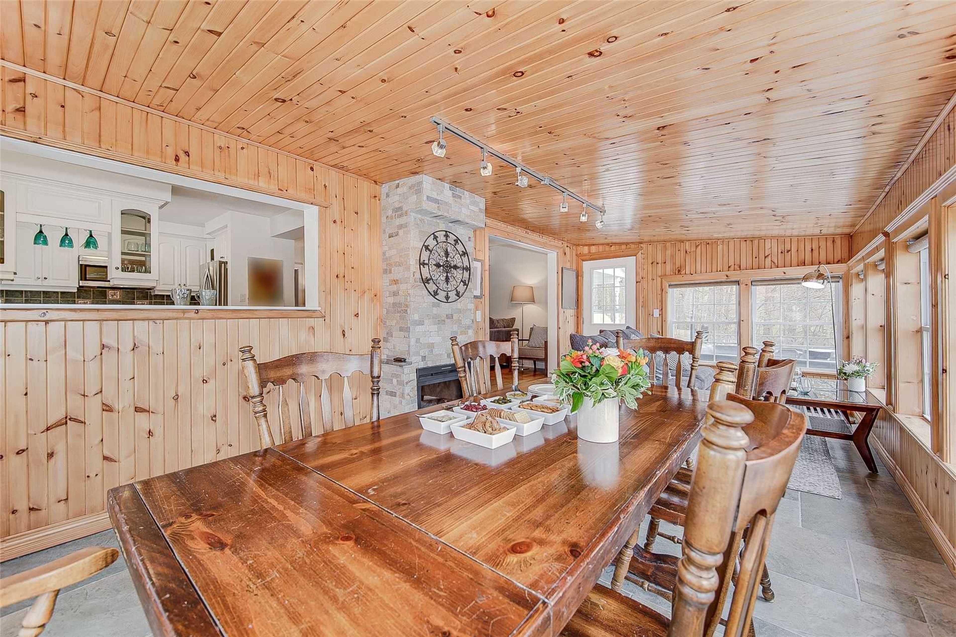 A large wooden dining table is set with snacks, surrounded by chairs in a room with wooden walls and ceiling, featuring a stone fireplace and large windows.