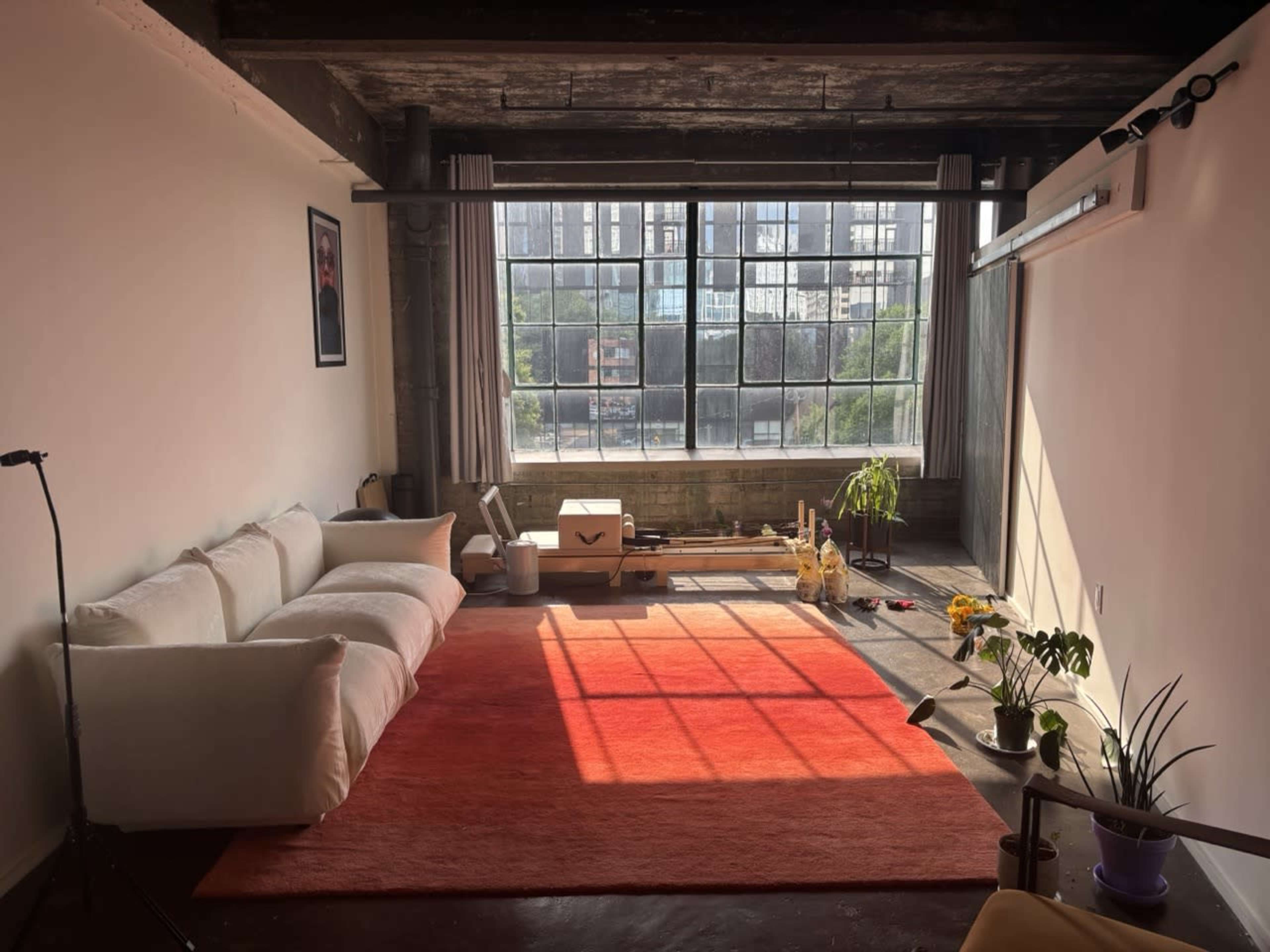 A spacious room with a large window, wooden beams, a white couch, orange rug, and plants near the sunlight.