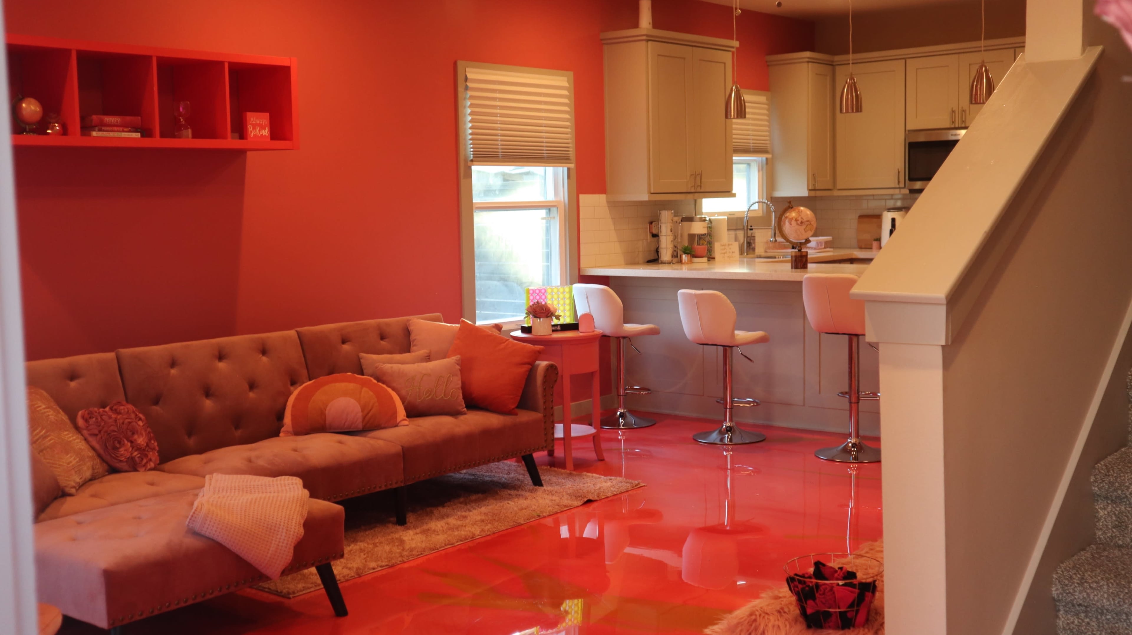 A bright, modern living space features a tufted sofa, colorful cushions, and a kitchen with bar stools and sleek cabinets, all against vibrant orange walls and a glossy floor.