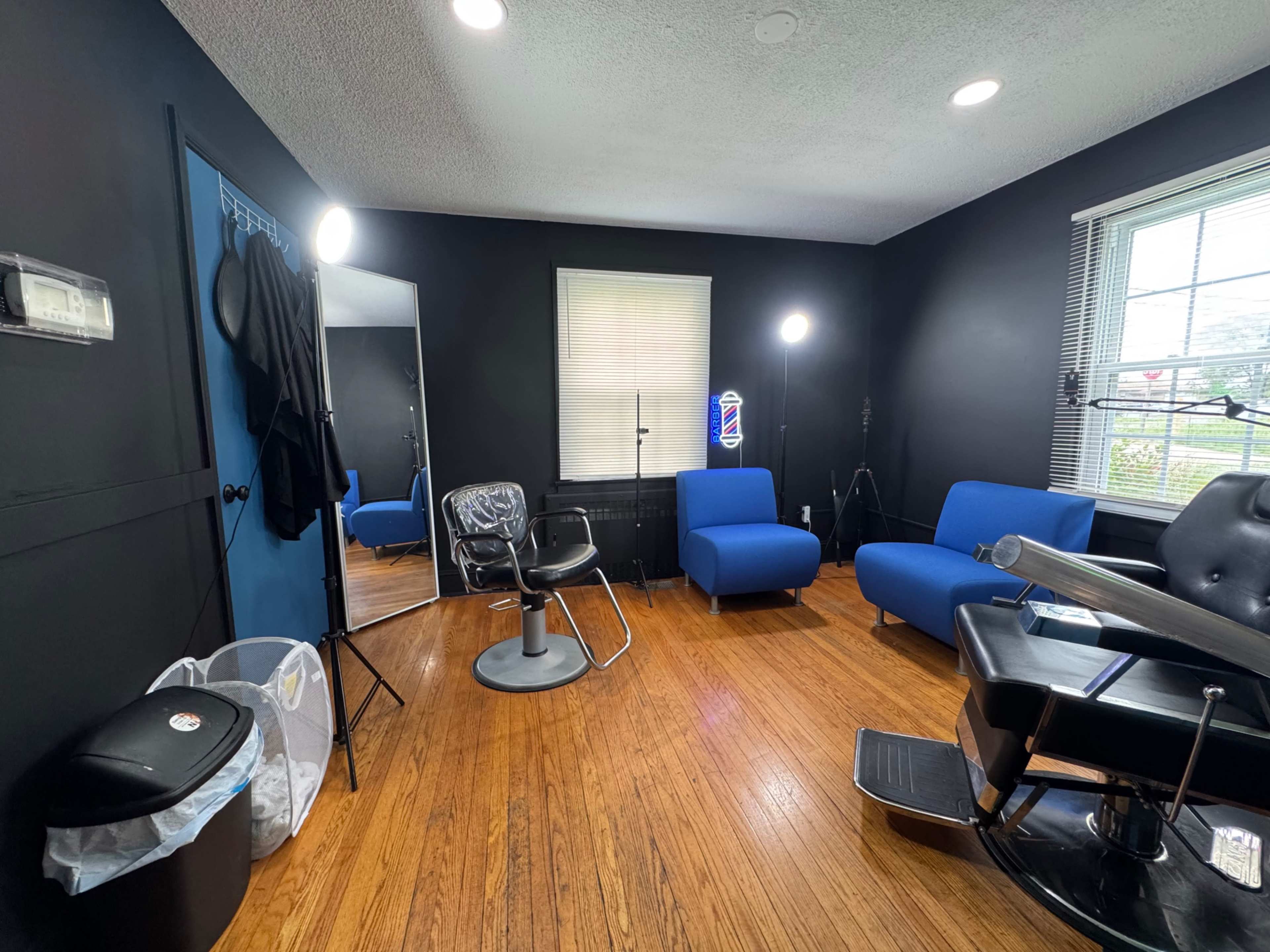 The image shows a barbershop interior with black walls, a chair for haircuts, a mirror, and two blue chairs.