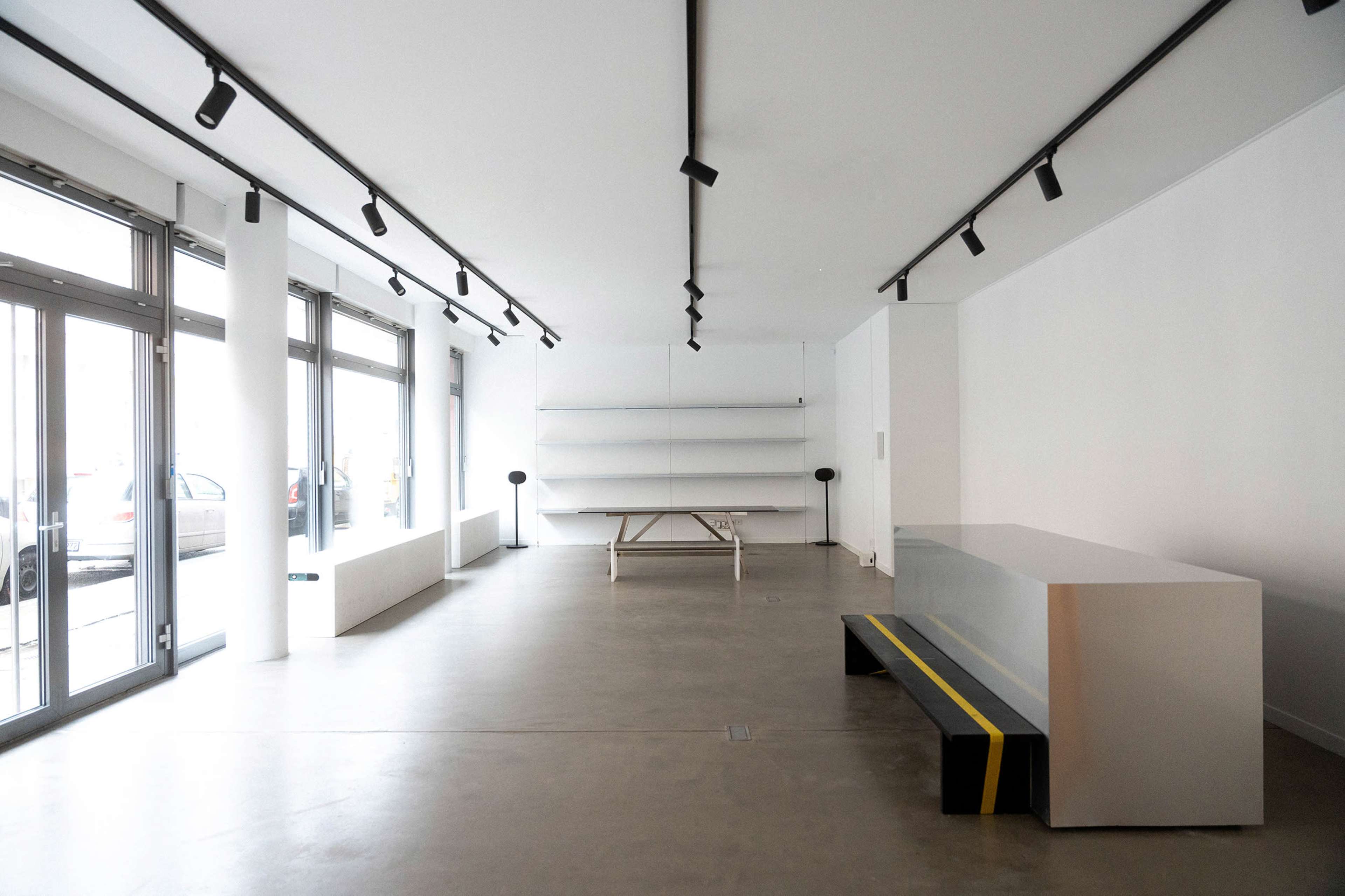 The image shows a minimalist interior space with white walls, a long wooden table, a few shelves, and seating that features a yellow stripe.