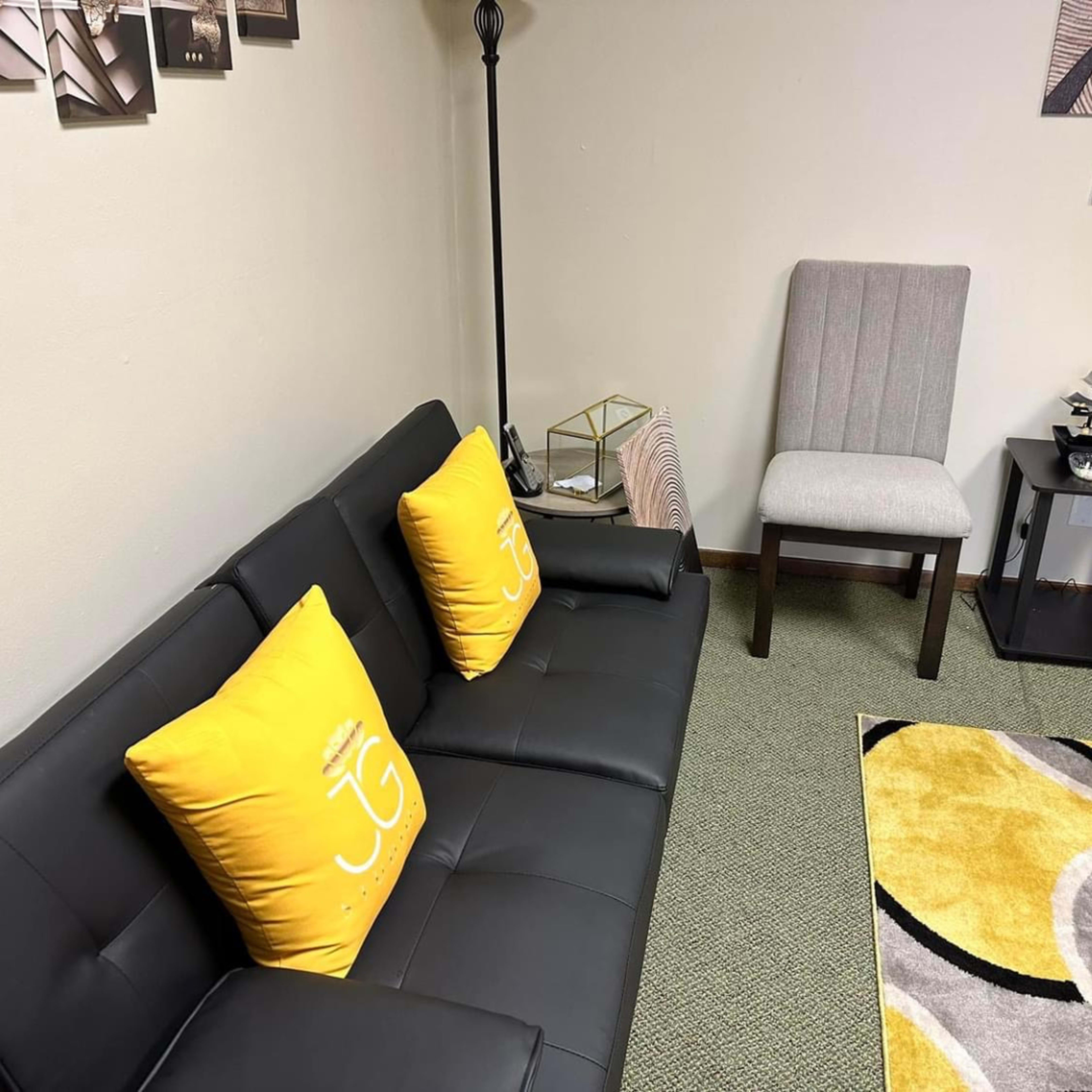 A black couch with two yellow cushions featuring a logo, alongside a light gray chair and a geometric-patterned rug on a green carpeted floor.