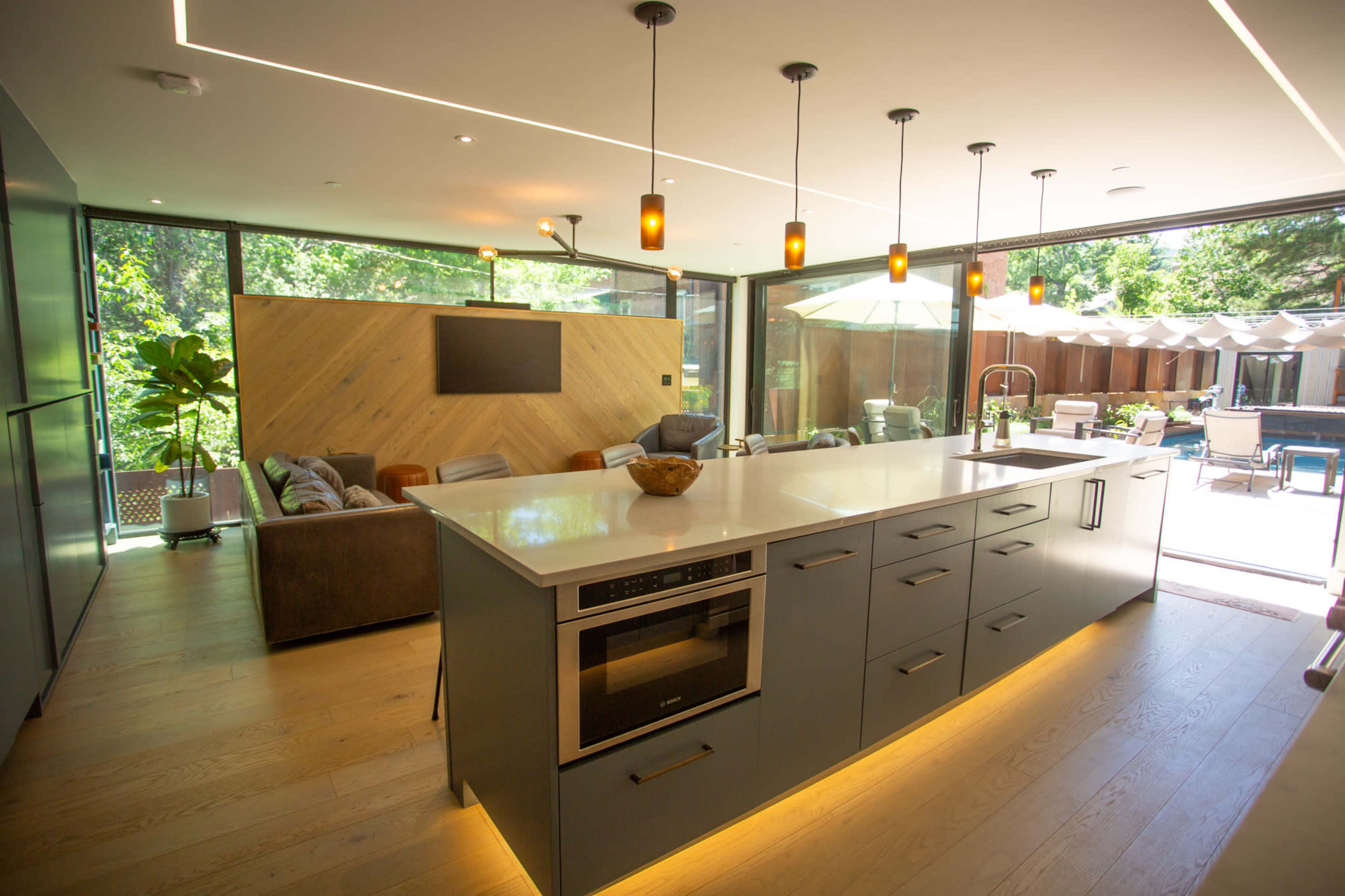 The image shows a modern kitchen with a central island, sleek cabinetry, and large windows that overlook a patio area with outdoor seating.
