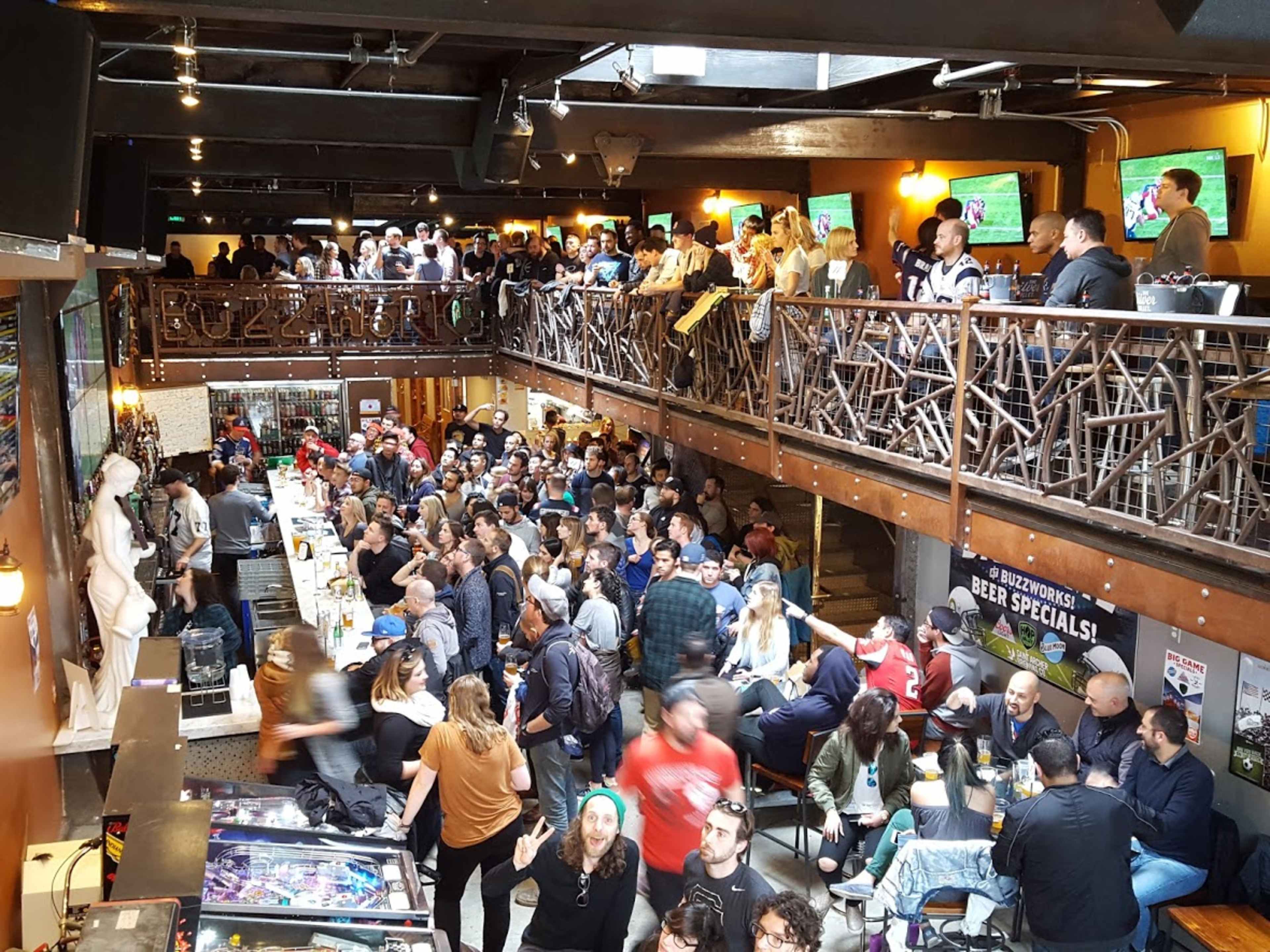 A crowded bar with multiple levels is filled with patrons watching sports on large screens and socializing.