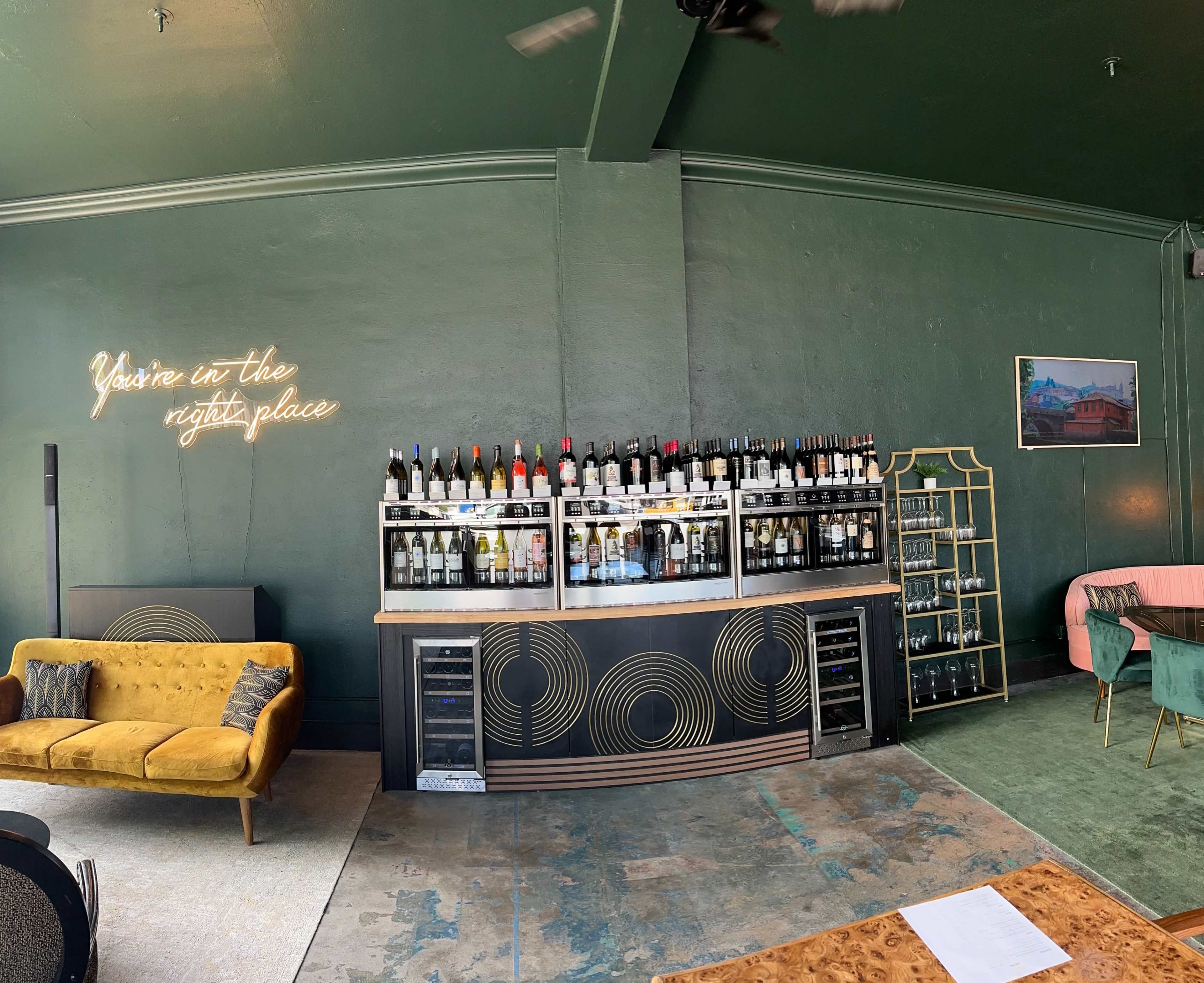 The image shows a modern bar area with a green wall, a wine shelf displaying various bottles, and stylish furniture, including a yellow couch and a pink chair.