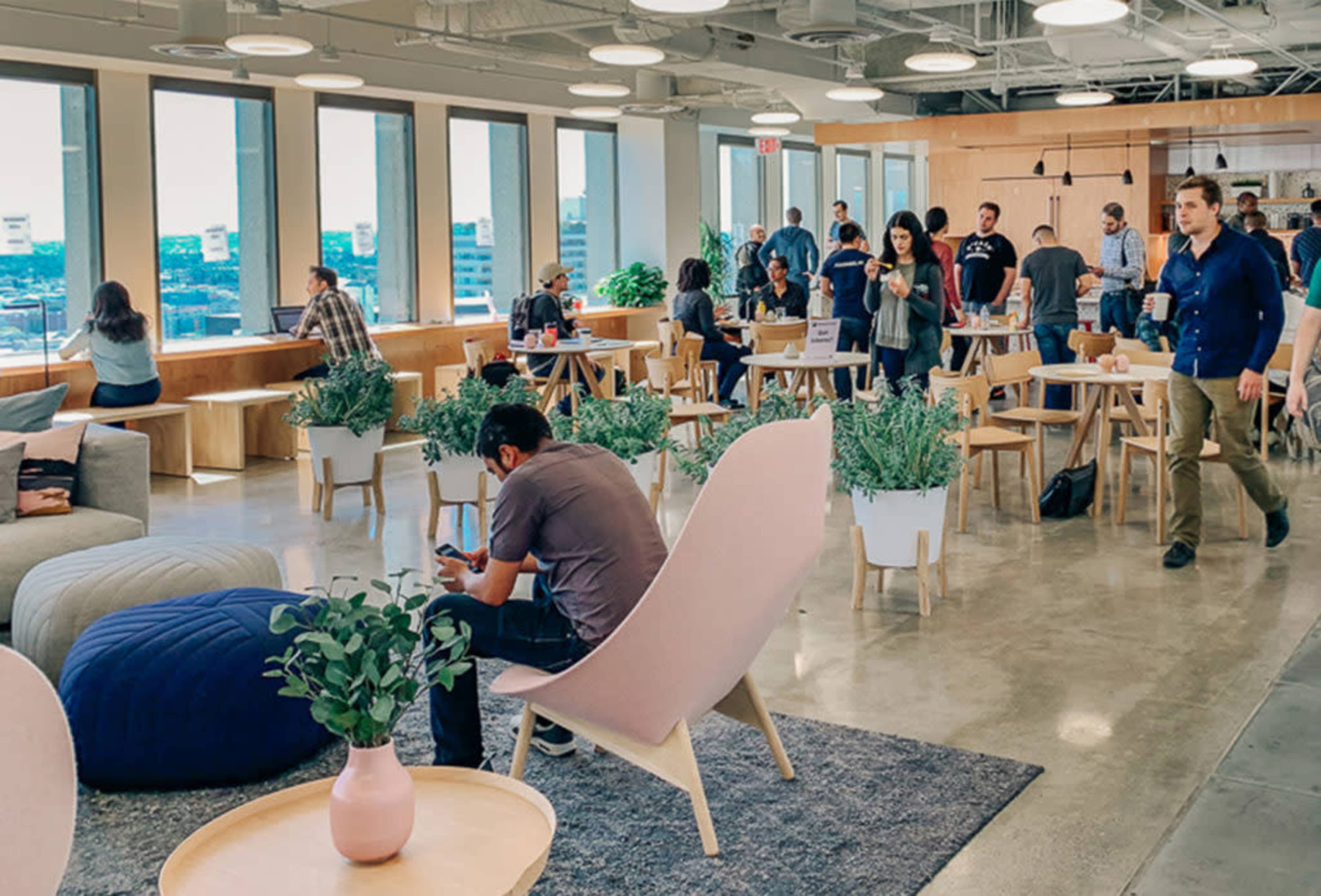 A modern office space features open seating areas, plants, and groups of people engaging in conversation and work activities.