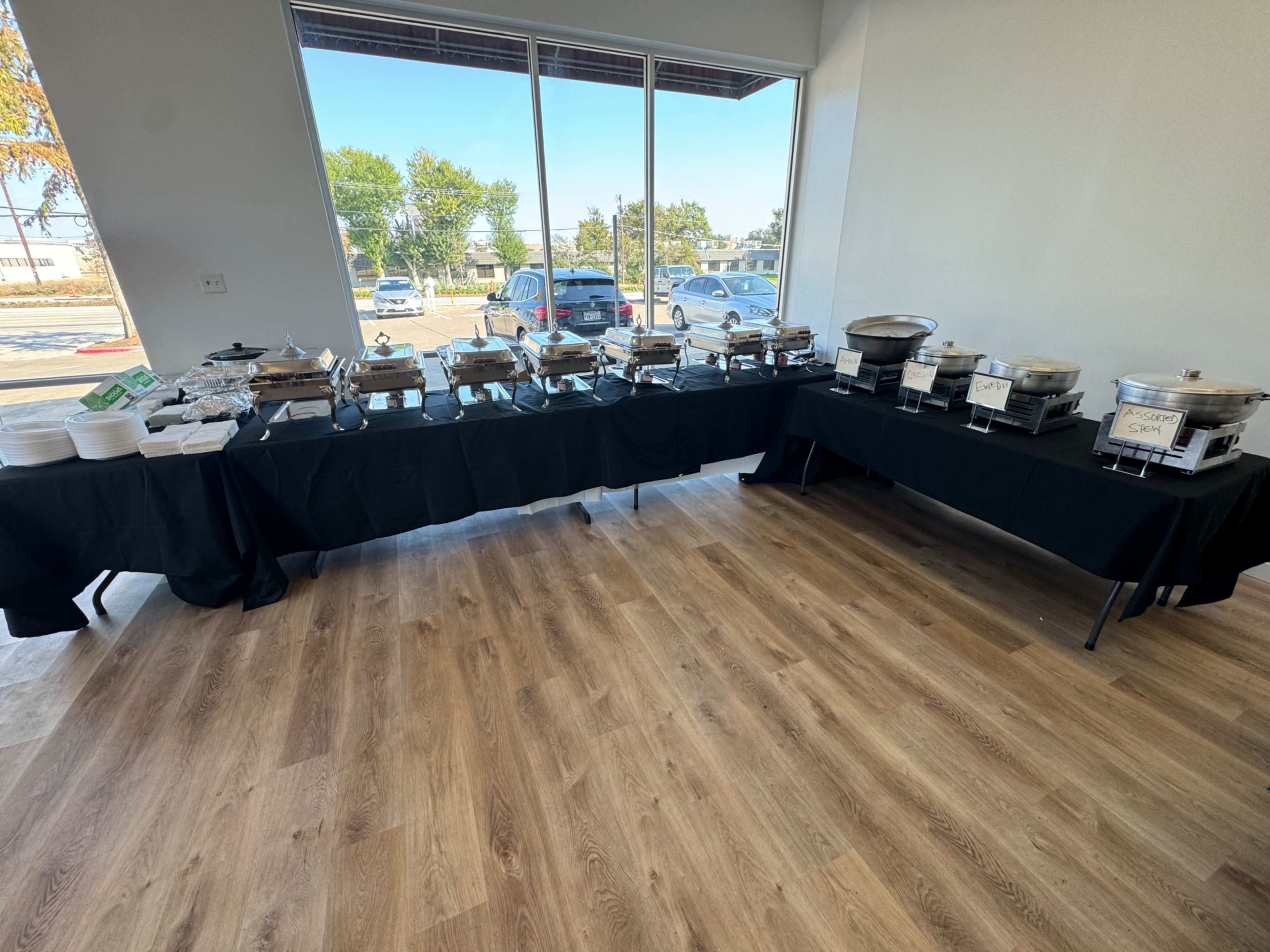 A spacious room features two long tables covered with black cloths, displaying numerous chafing dishes and serving utensils, with large windows letting in natural light.