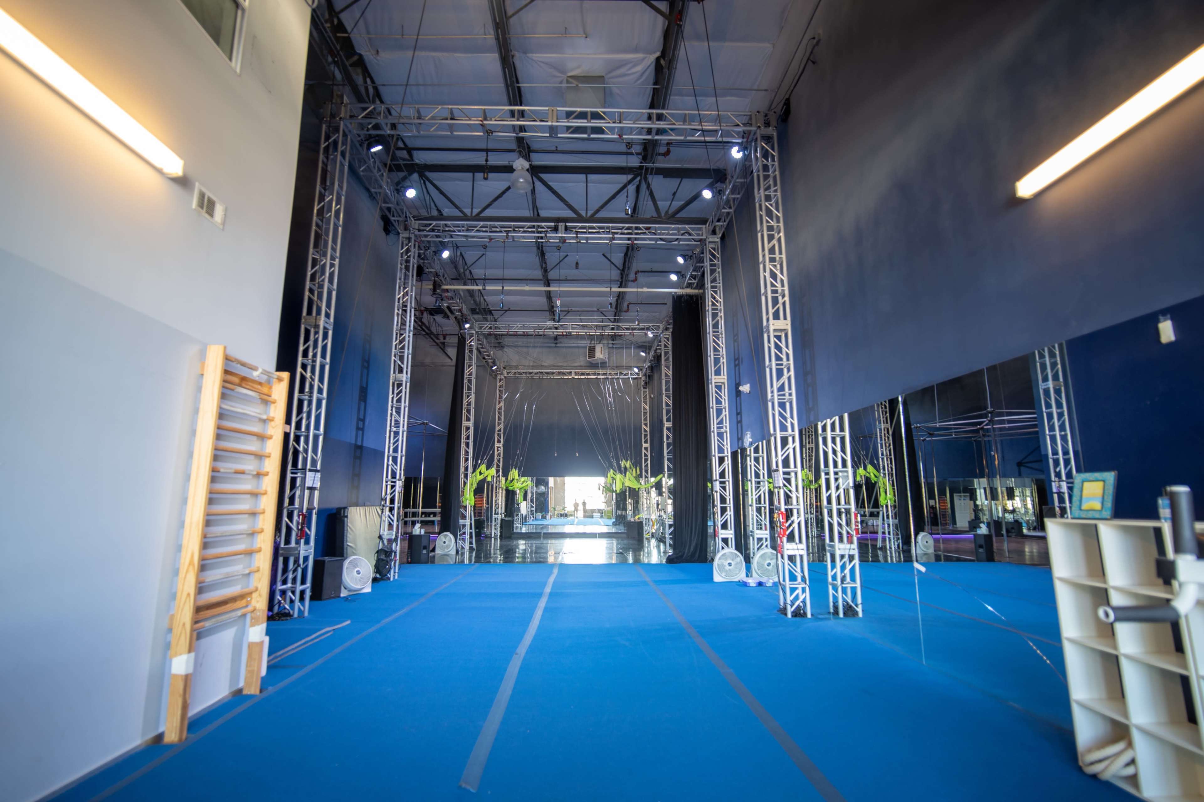 An expansive indoor gymnasium features blue flooring, mirrored walls, and lighting supported by metal trusses.