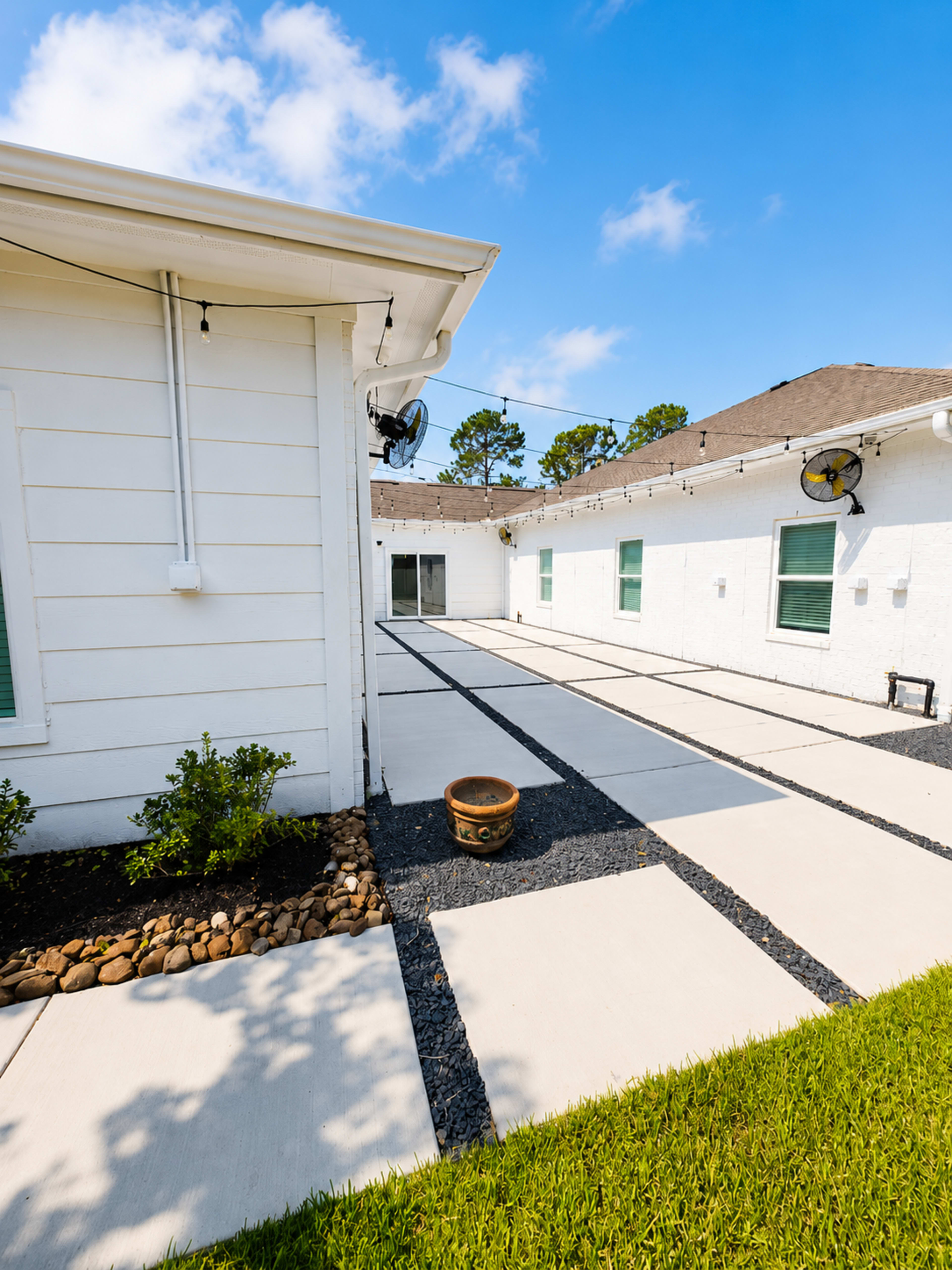 The image shows a clean, open courtyard with concrete pathways and decorative landscaping, bordered by white walls and a grassy area.