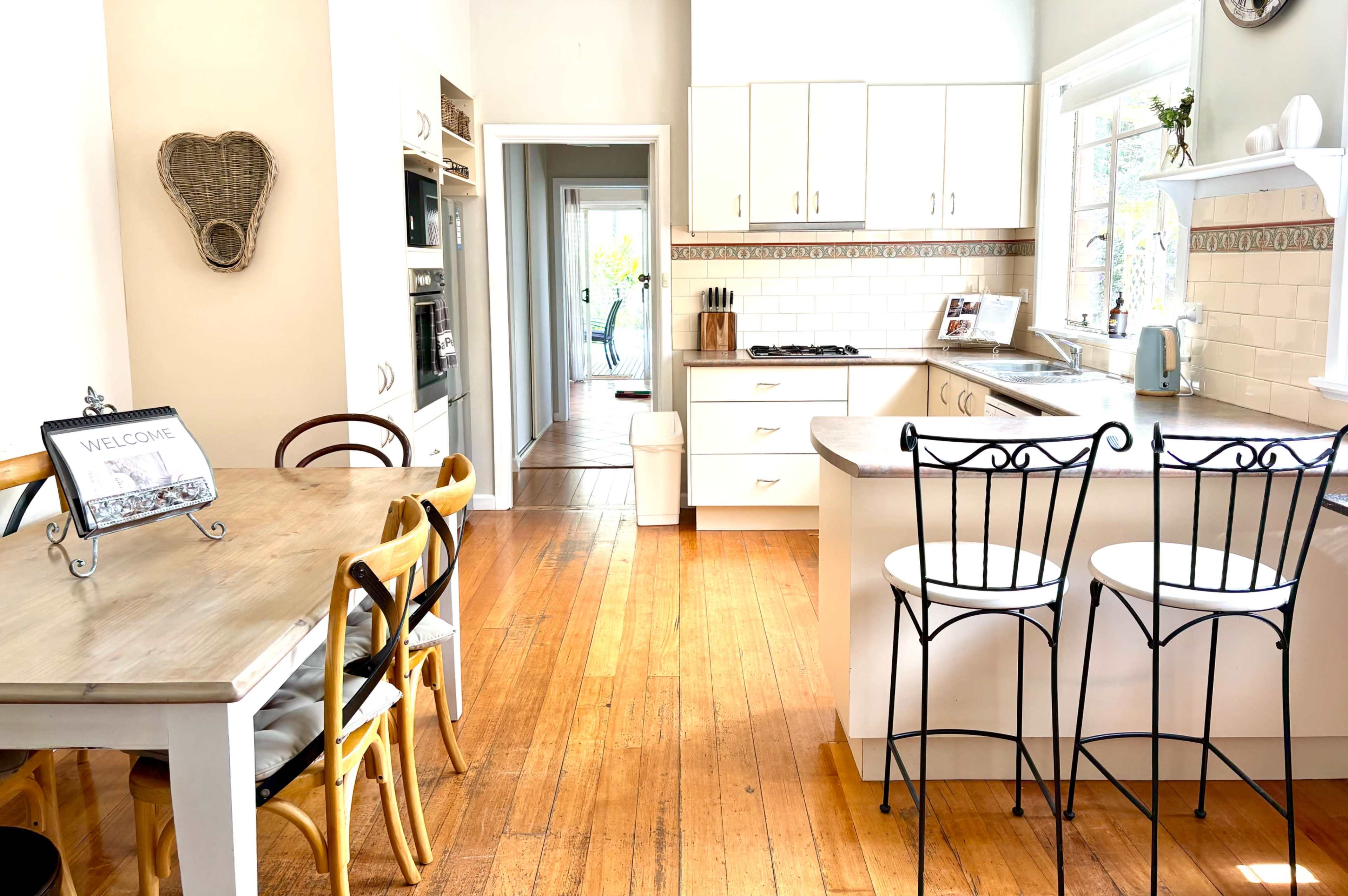 The image shows a bright and clean kitchen with wooden floors, equipped with modern appliances, a dining table with chairs, and a view leading to a hallway.