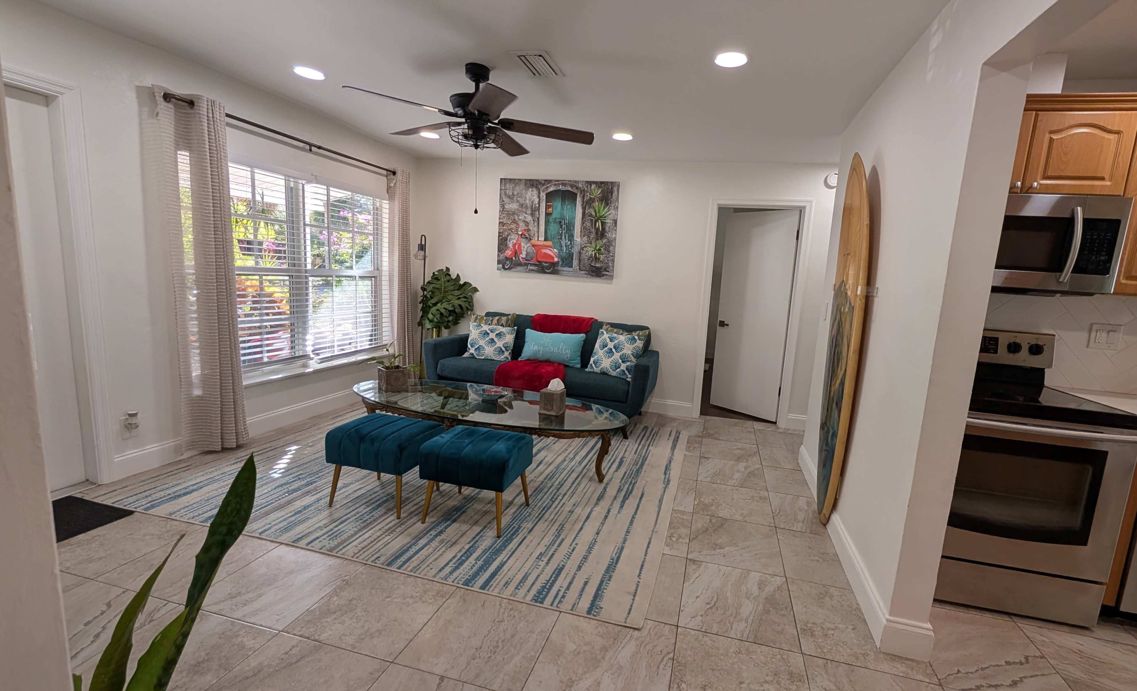 The image shows a modern living room with a teal sofa, a glass coffee table, and a patterned area rug, illuminated by natural light through a window.